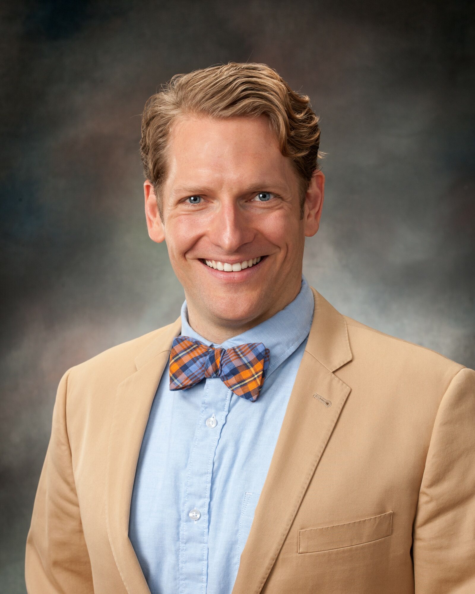 Portrait of a smiling man with light brown hair, wearing a beige blazer, light blue shirt, and a plaid bow tie.