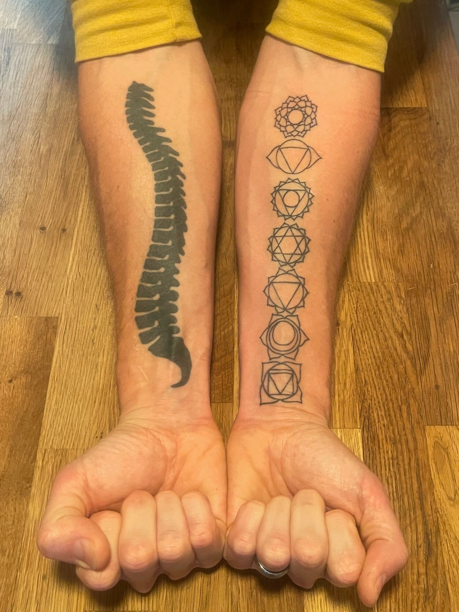 Two forearms with tattoos, one with a black spine tattoo and the other with geometric designs, resting on a wooden floor.