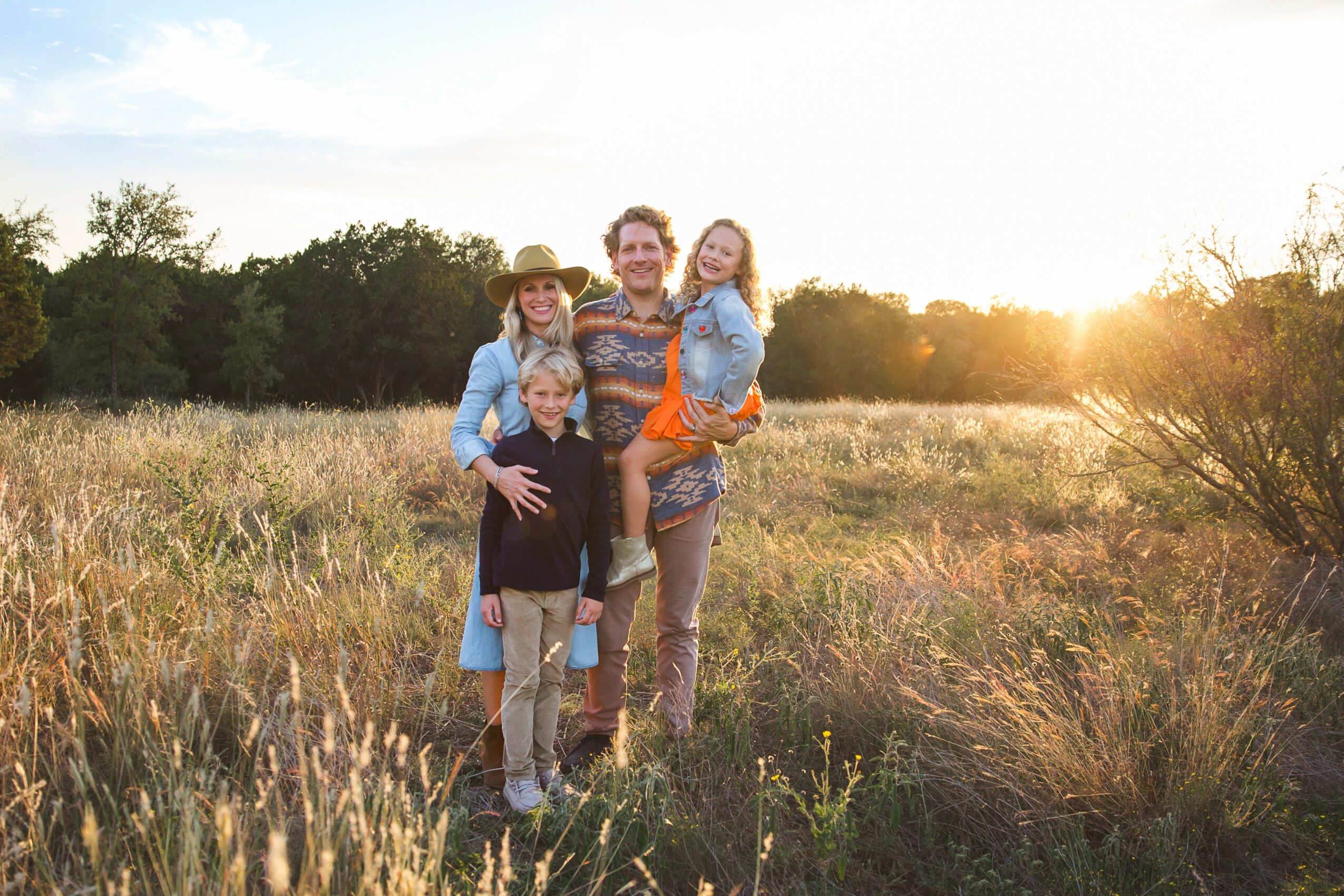 Family of four standing in a field at sunset, smiling, with trees in the background.