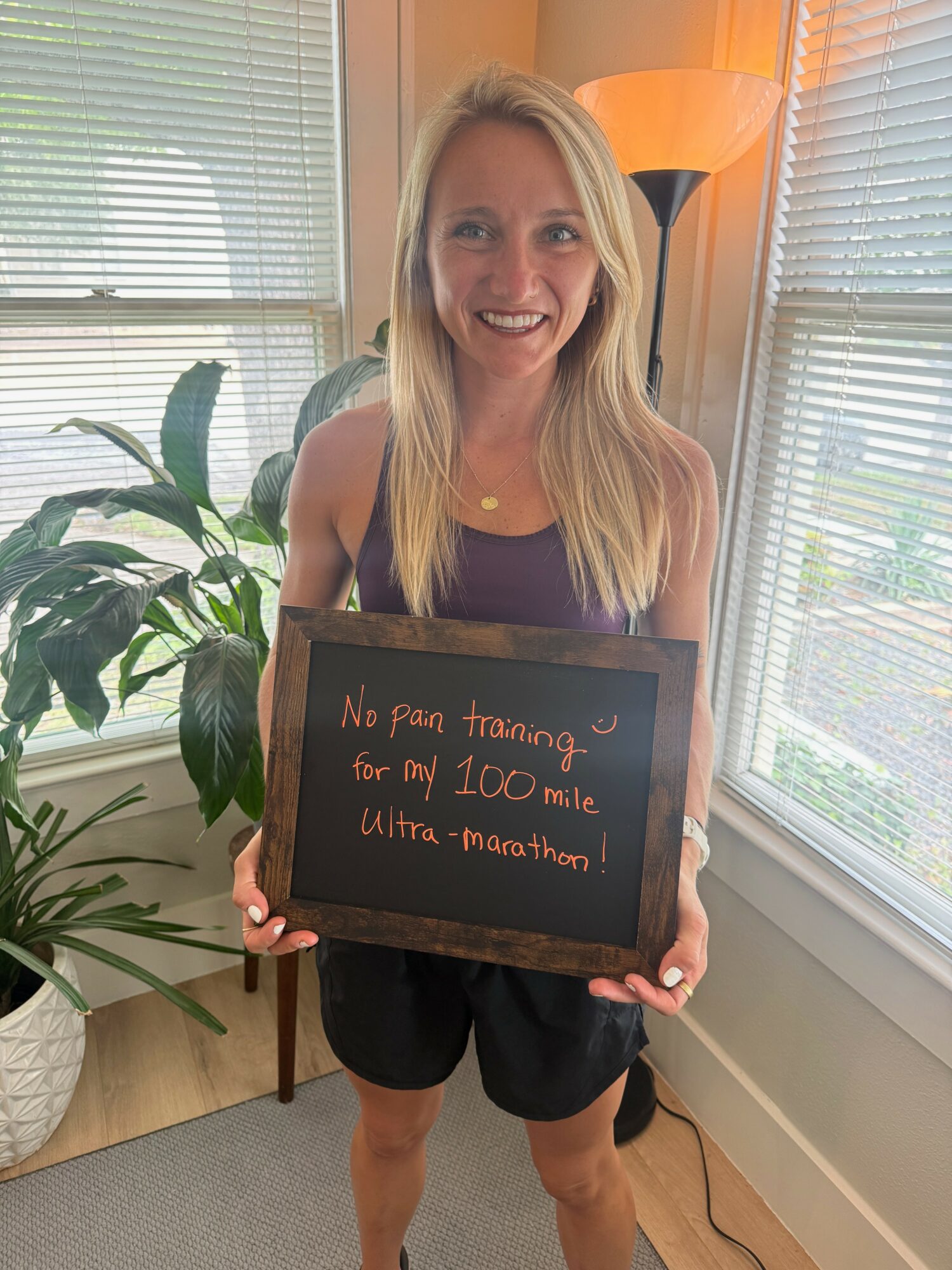 Smiling woman holding a chalkboard with handwritten message in a bright room with windows and a plant.