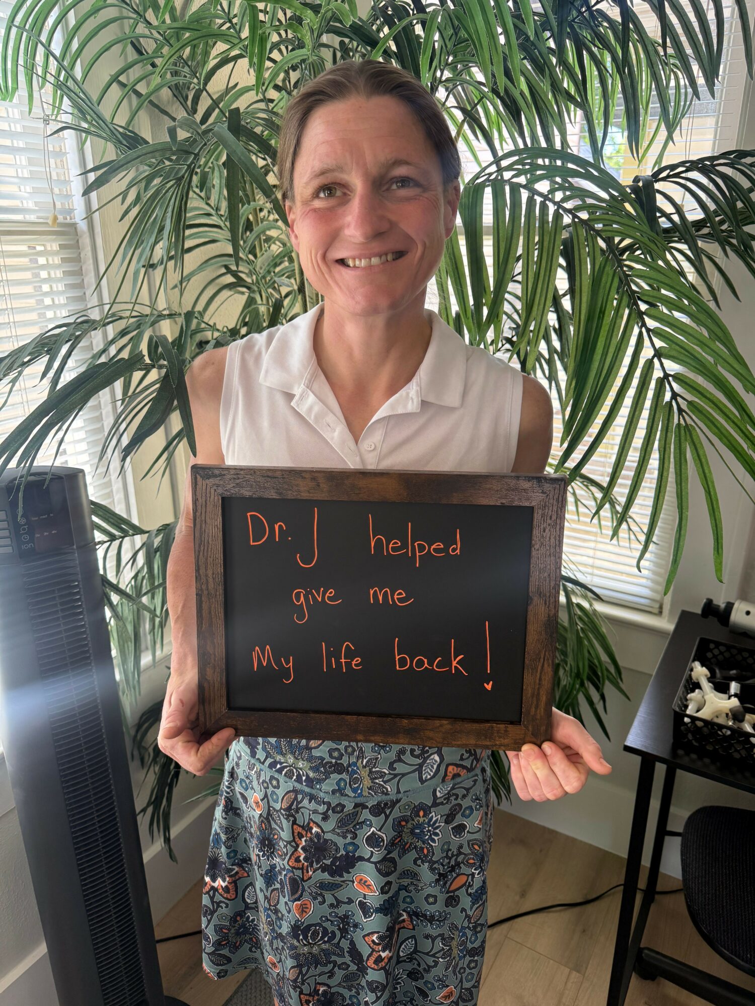 Woman holding a sign that says 'Dr J helped me when we were back!' in front of large green plants.