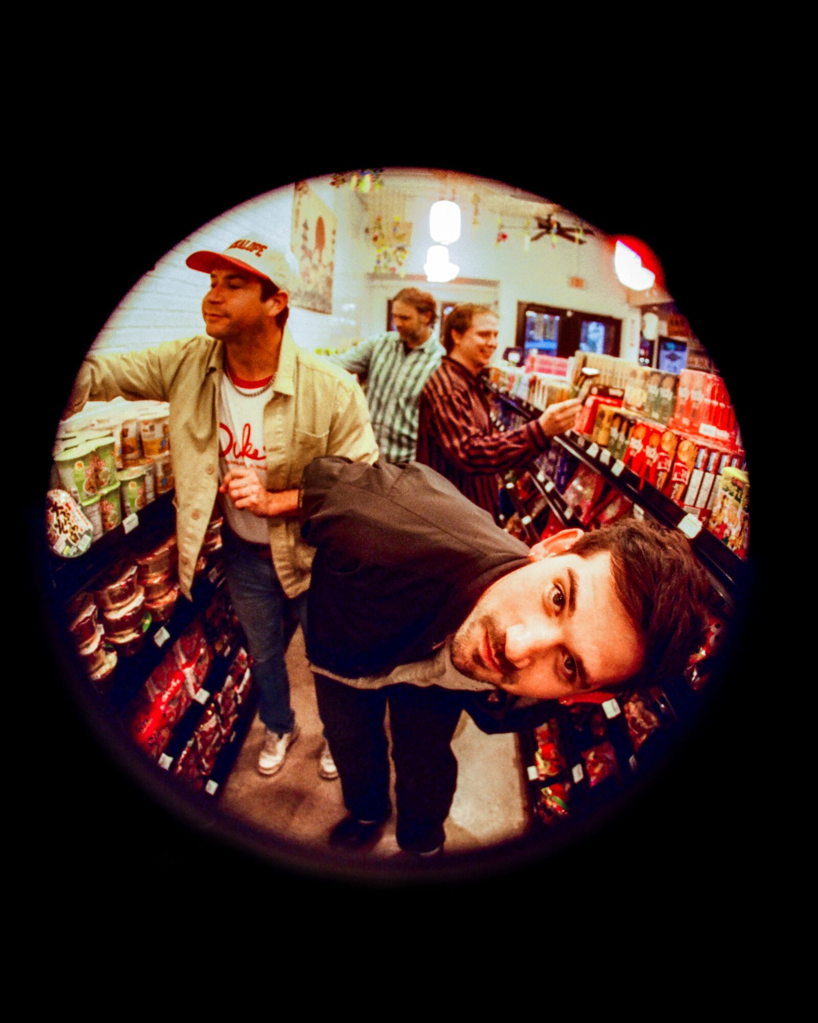 Group of people shopping in a store aisle with shelves of products, some looking at the camera, one leaning in close.