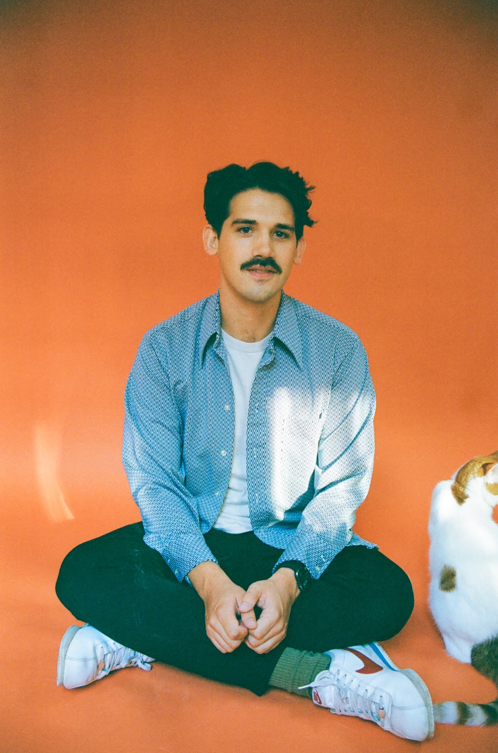 Young man with dark hair and mustache sitting cross-legged against an orange background, wearing a blue shirt and white sneakers.