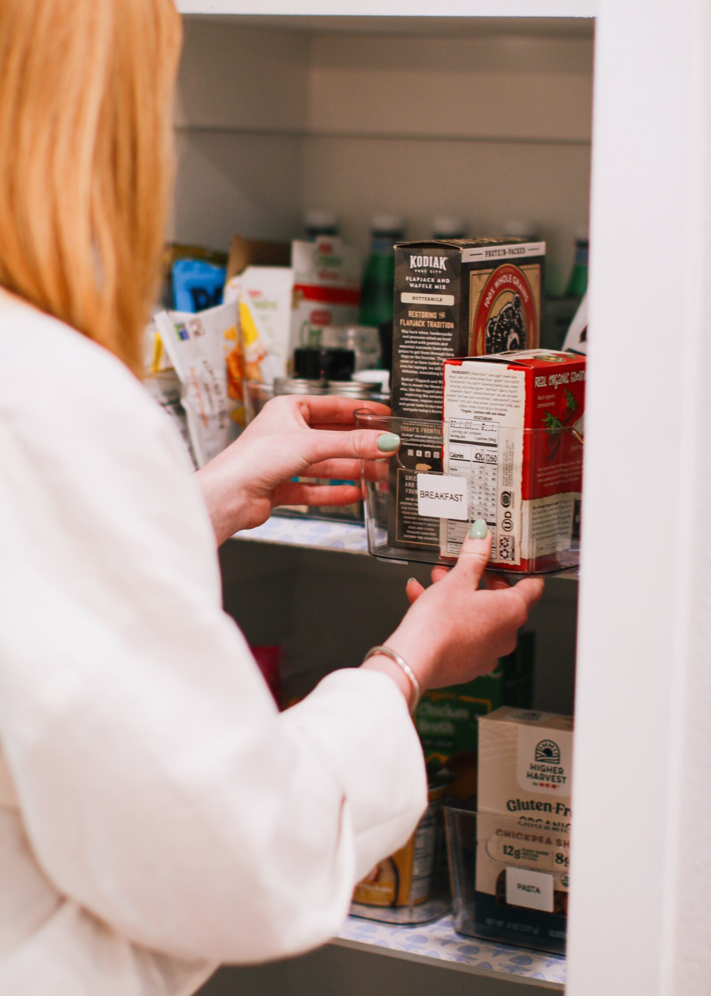 Person with red hair reaching into a pantry shelf filled with canned and boxed food items.