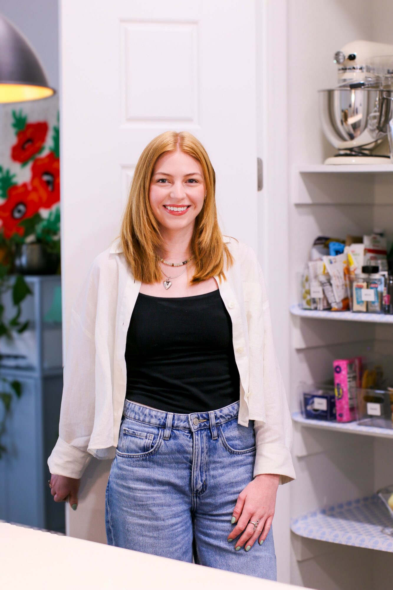 Young woman with red hair smiling in a kitchen, wearing a black top, light blue jeans, and a white cardigan.