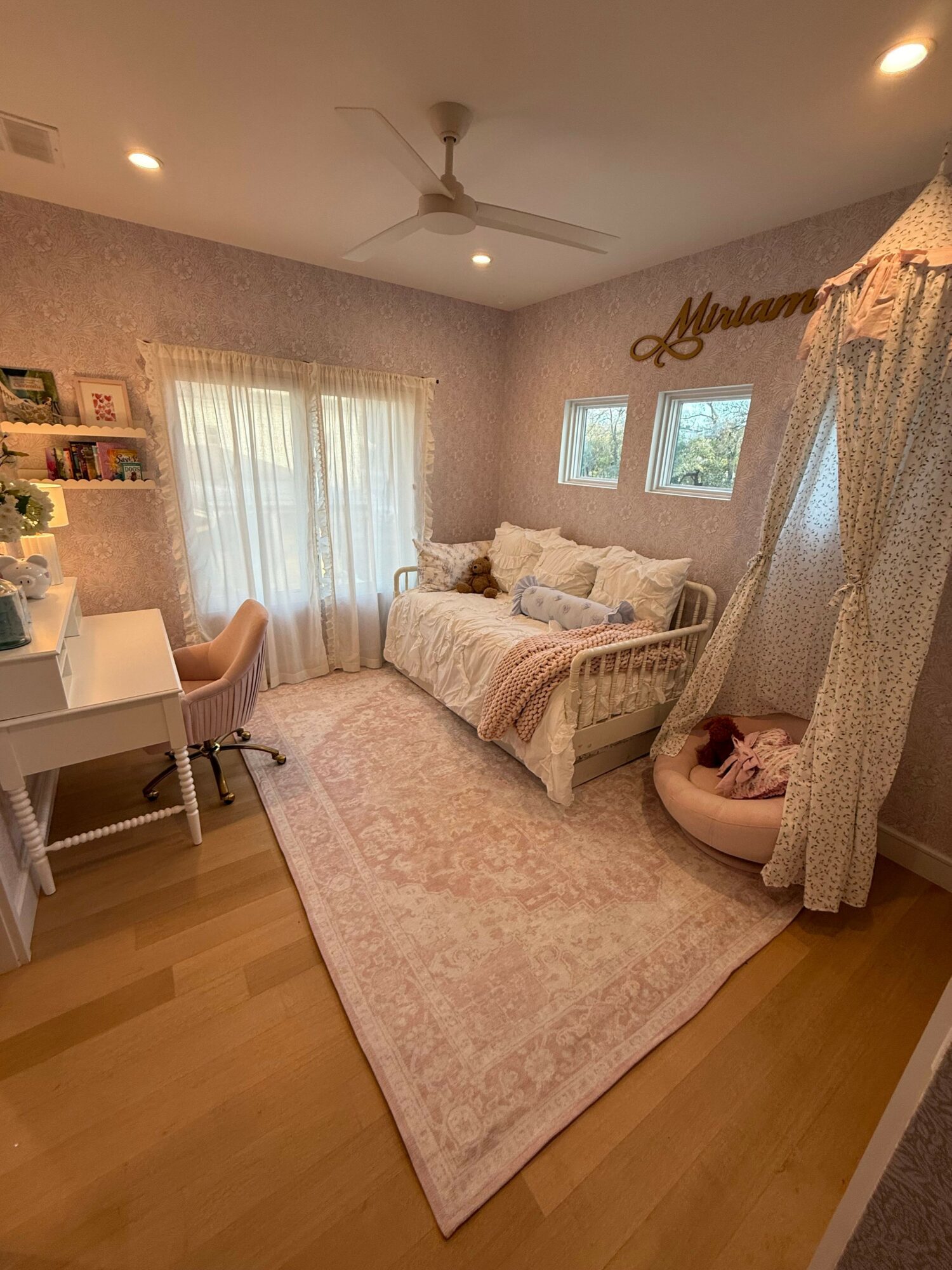 Child lying on bed in a cozy bedroom with pink and white decor, curtains, and a pink rug.