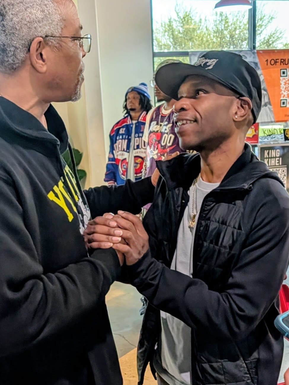 Two men shake hands and smile at each other indoors, with a woman and a poster in the background.