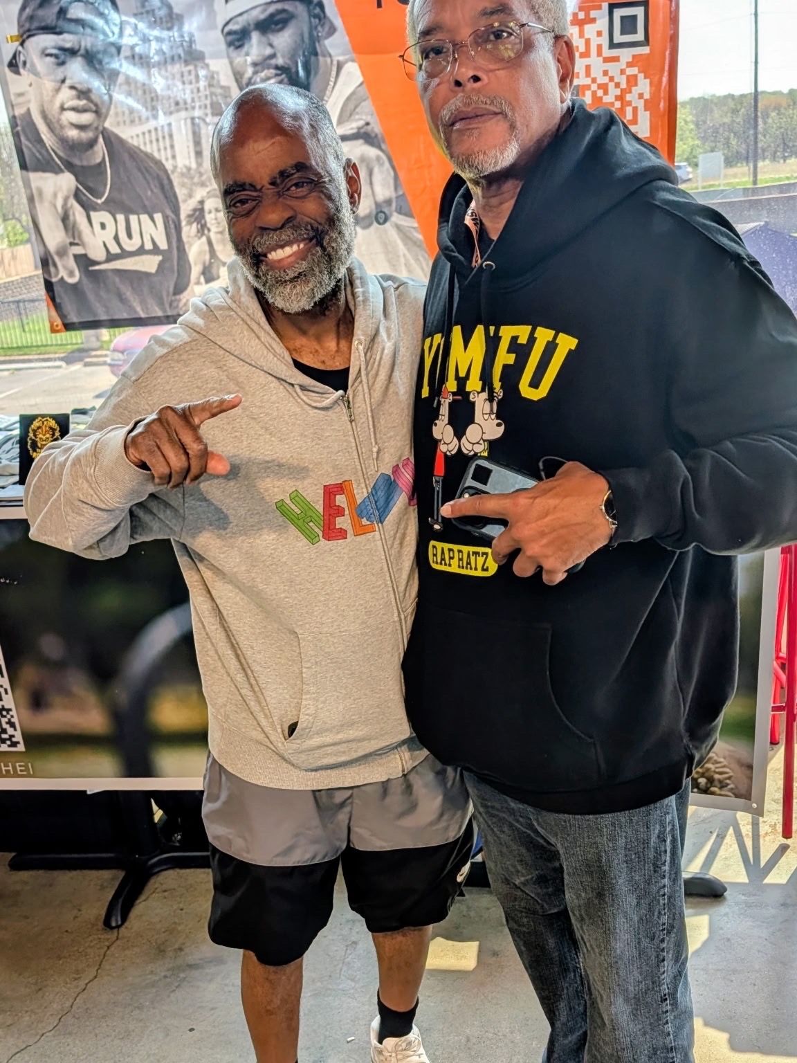 Two men smiling indoors, one pointing, the other holding a phone, with posters in the background.
