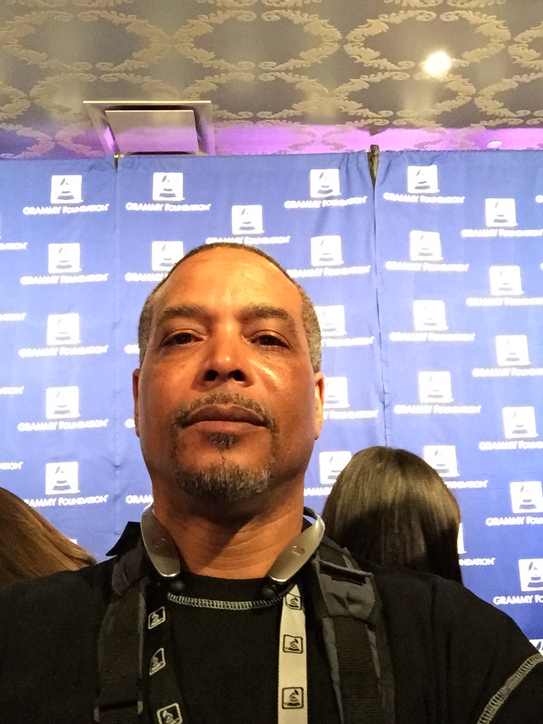 Man with short hair and a beard taking a selfie at an event with a blue backdrop and people in the background.