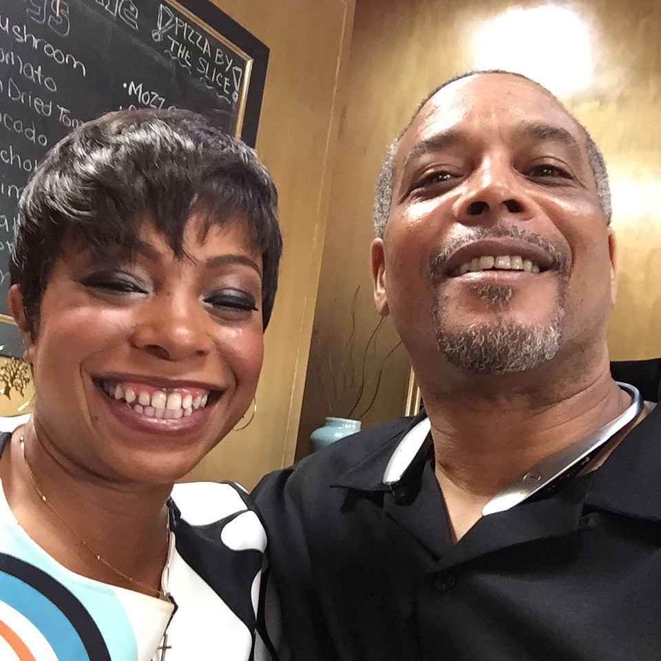 Two people smiling, one with short dark hair and the other with a shaved head, in a restaurant setting.