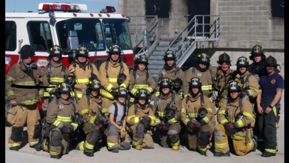 Group of firefighters in uniform and helmets, standing outdoors with fire truck in background.