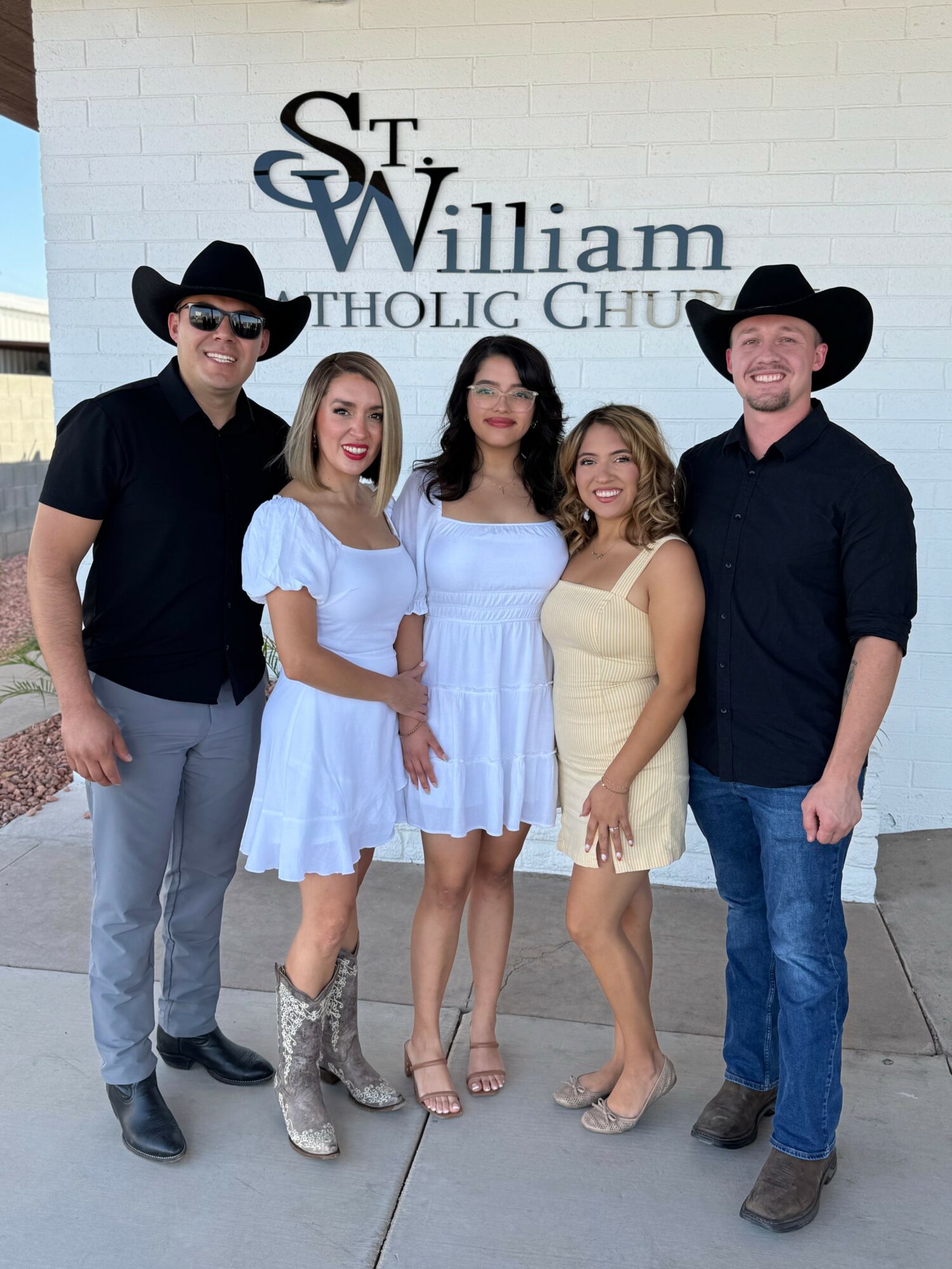 Group of five people standing outside in front of a white brick wall with a sign, smiling, dressed in casual and cowboy attire.