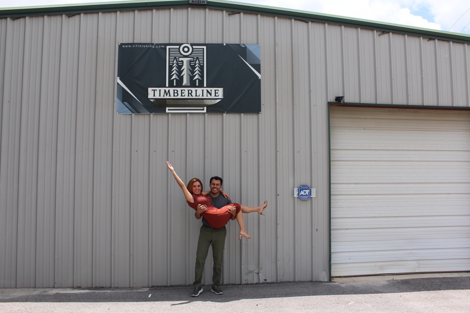 Two people standing in front of a gray industrial building with a Timberline sign, one person pointing upward, both smiling.