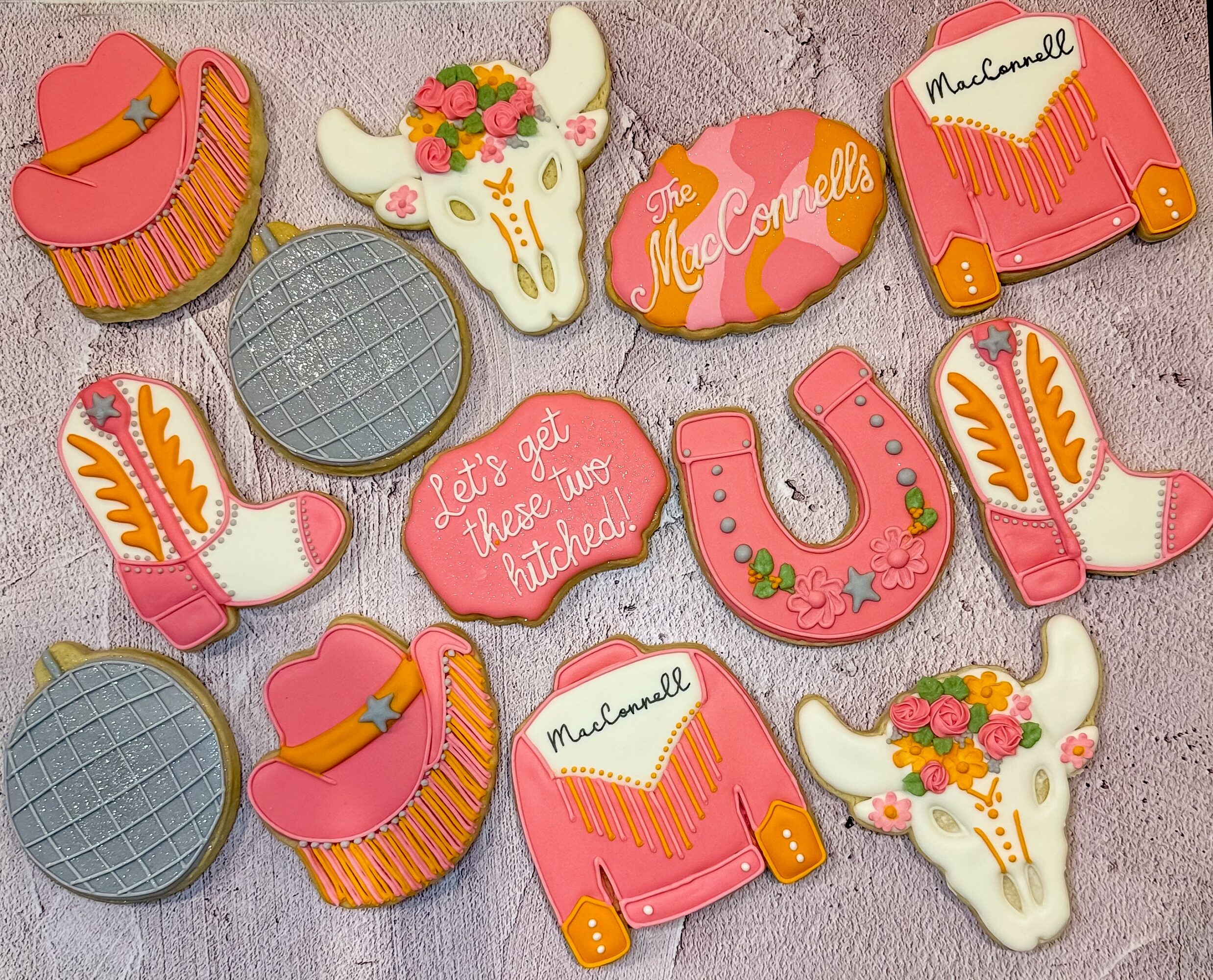 Decorated cookies with pink, orange, and white colors, including a cupcake, cow skull, heart, boots, and horseshoe, on a pink surface.