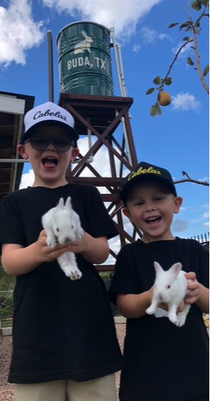 Two children holding white rabbits outdoors, water tower in background, blue sky, trees, and clouds.