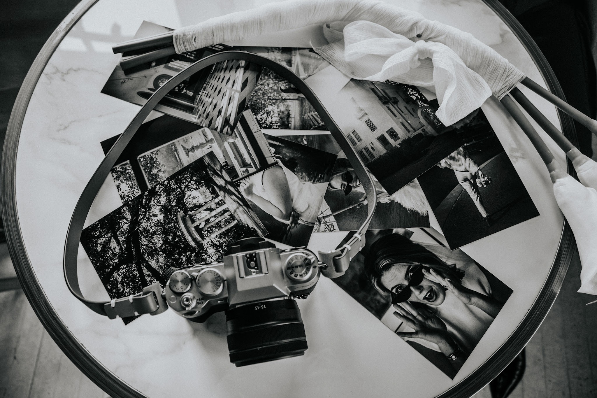 A camera, photographs, and a strap are arranged on a glass table with a white cloth and some objects nearby.