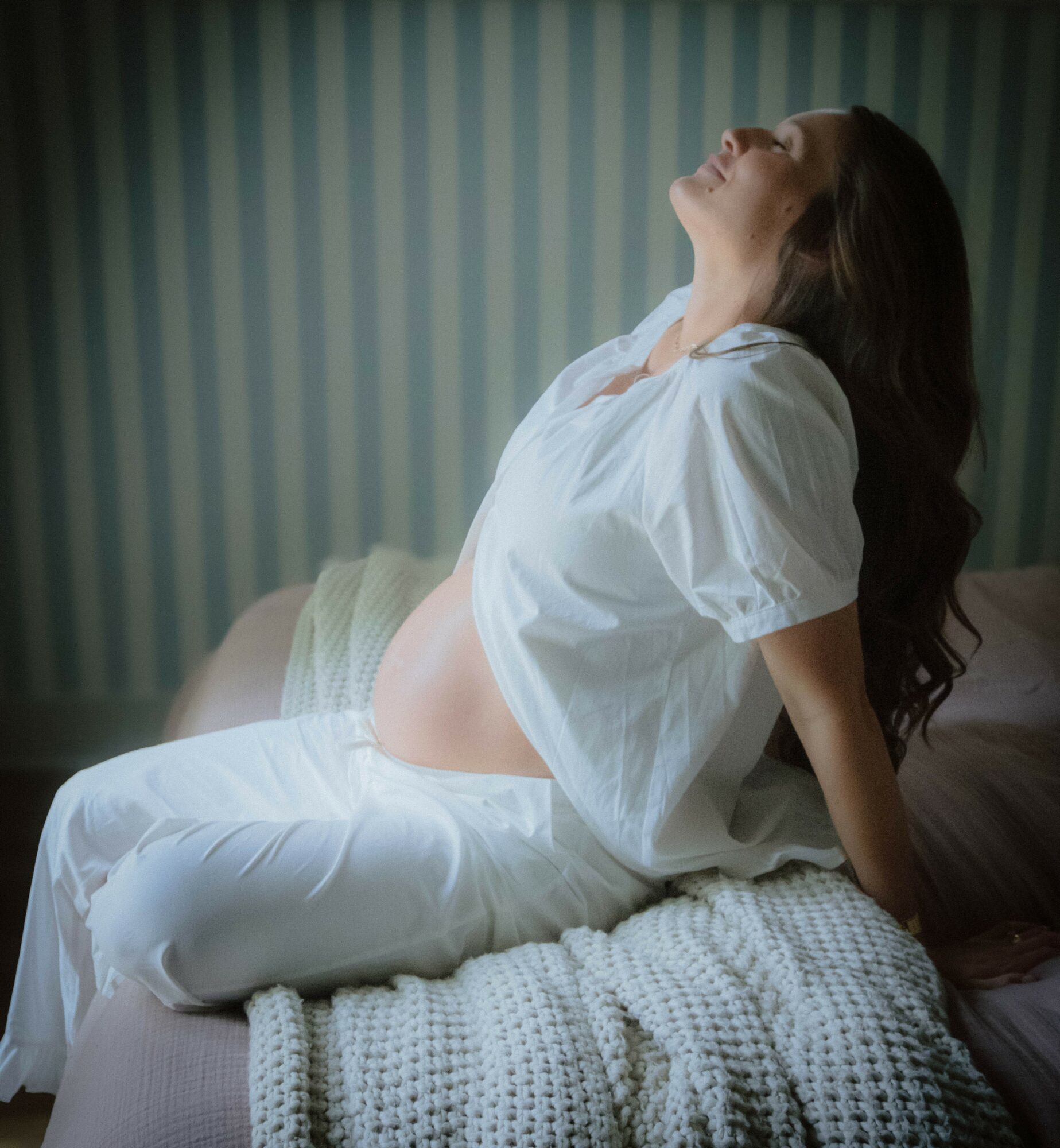Pregnant woman sitting on bed, leaning back with head tilted upward, in a room with striped wallpaper.