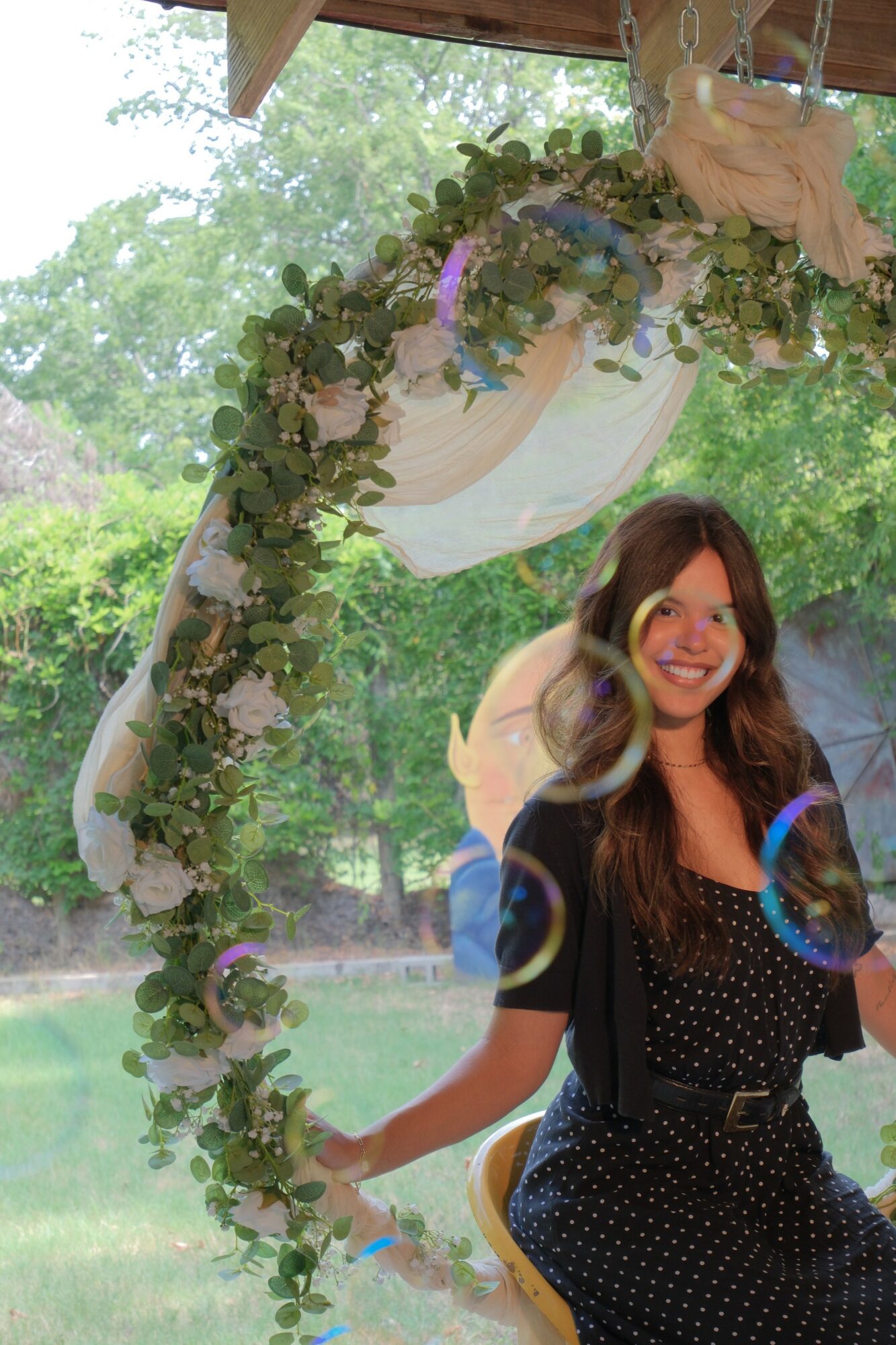 Young woman with long hair smiling outdoors, surrounded by greenery and trees, under a decorated arch with flowers and fabric.