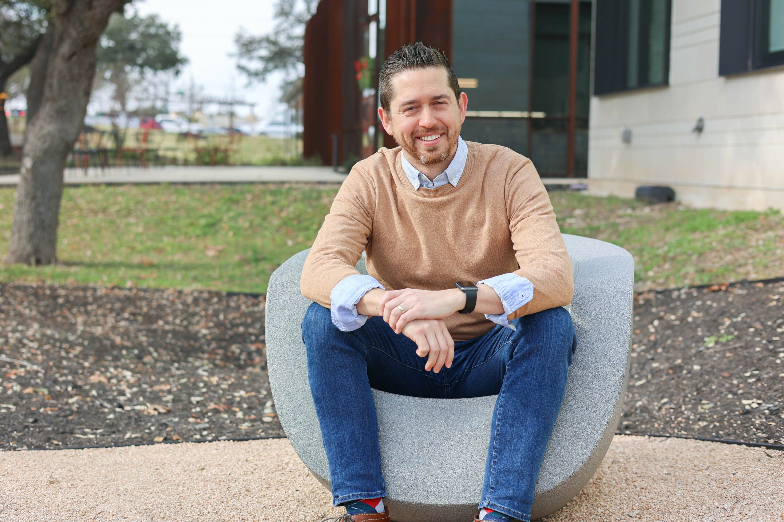 Smiling man sitting outdoors on a modern gray chair, wearing a tan sweater and blue jeans, with a building and trees in background.