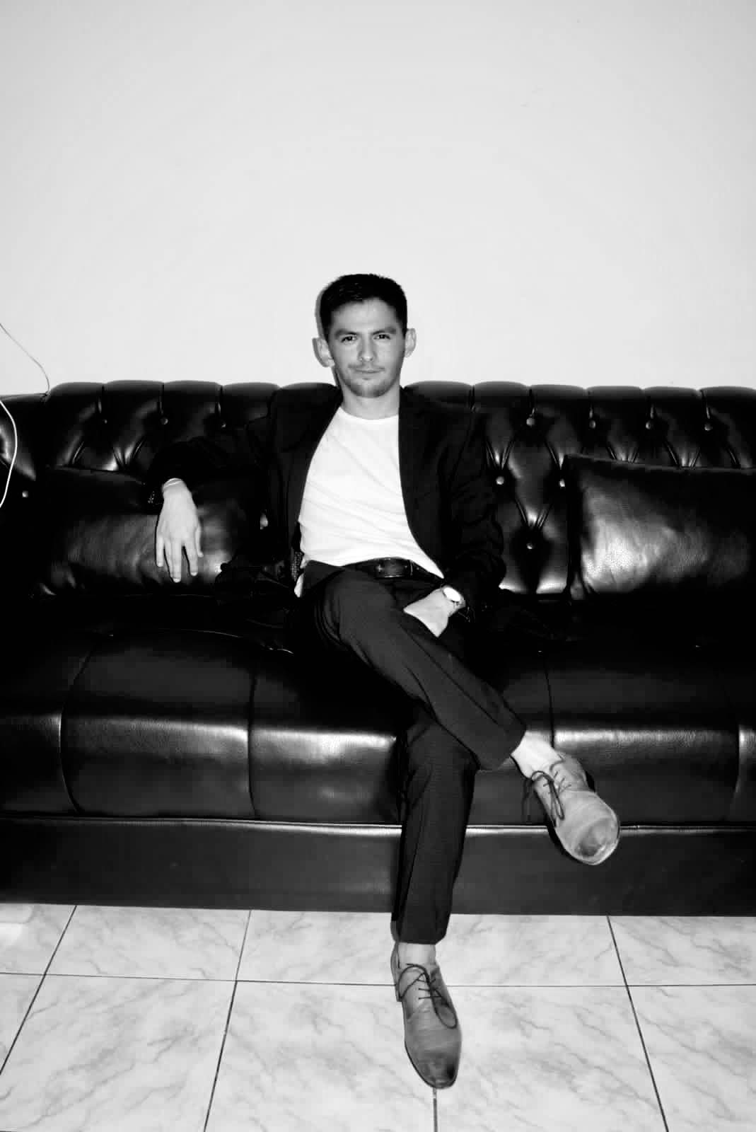 Man sitting on a black leather couch with crossed legs, wearing a blazer and white shirt, in a minimal room.