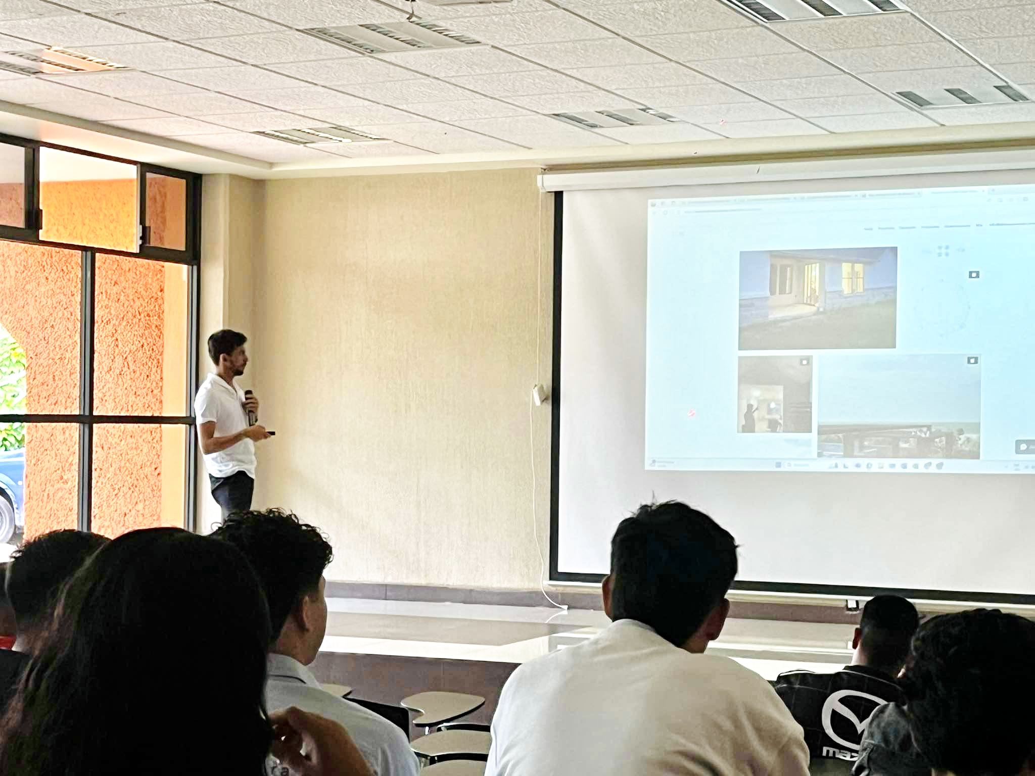 Person presenting in front of a projection screen with audience seated, in a room with large windows.