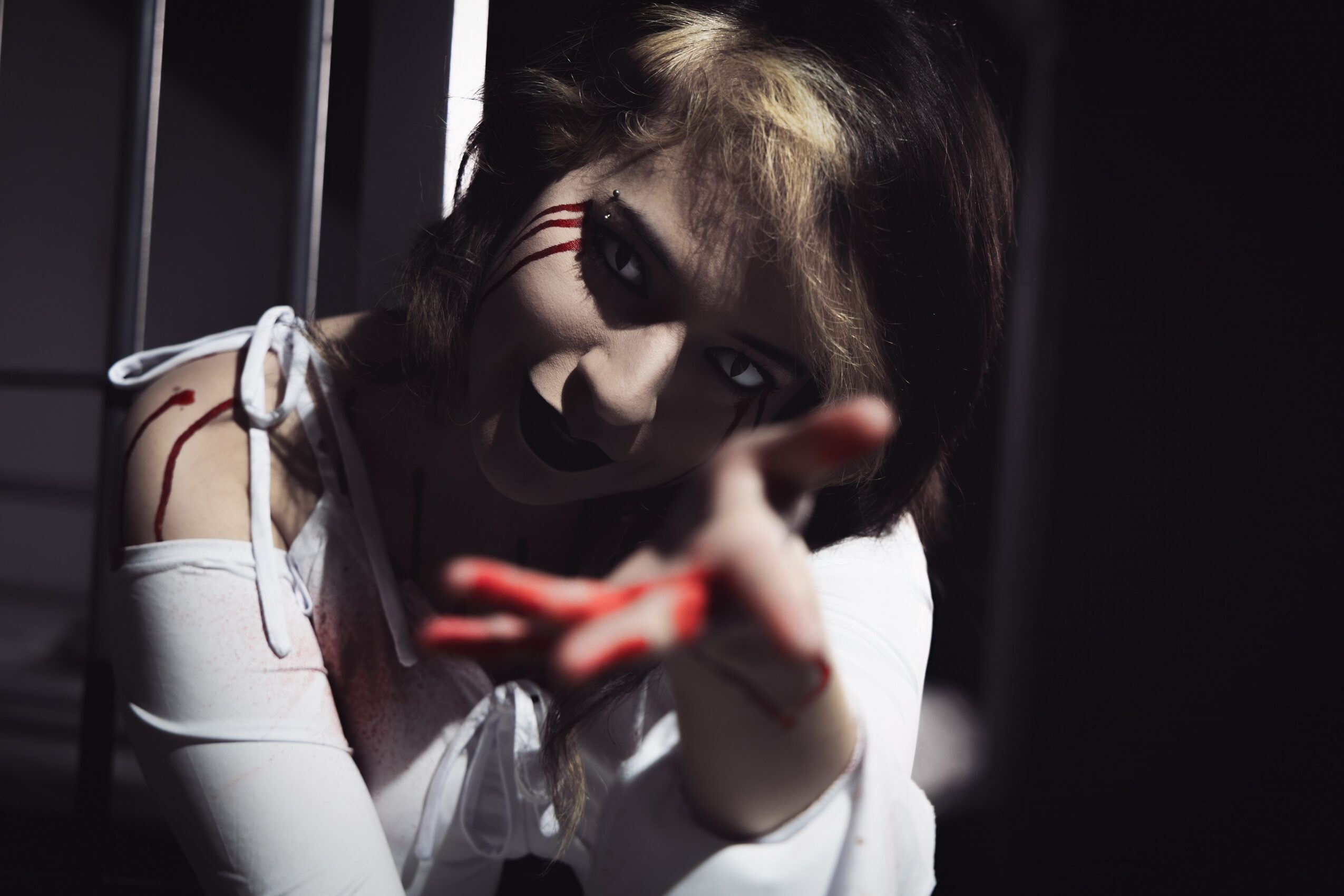 Person with dark makeup and long hair reaching toward the camera, behind metal bars, in a dark setting.
