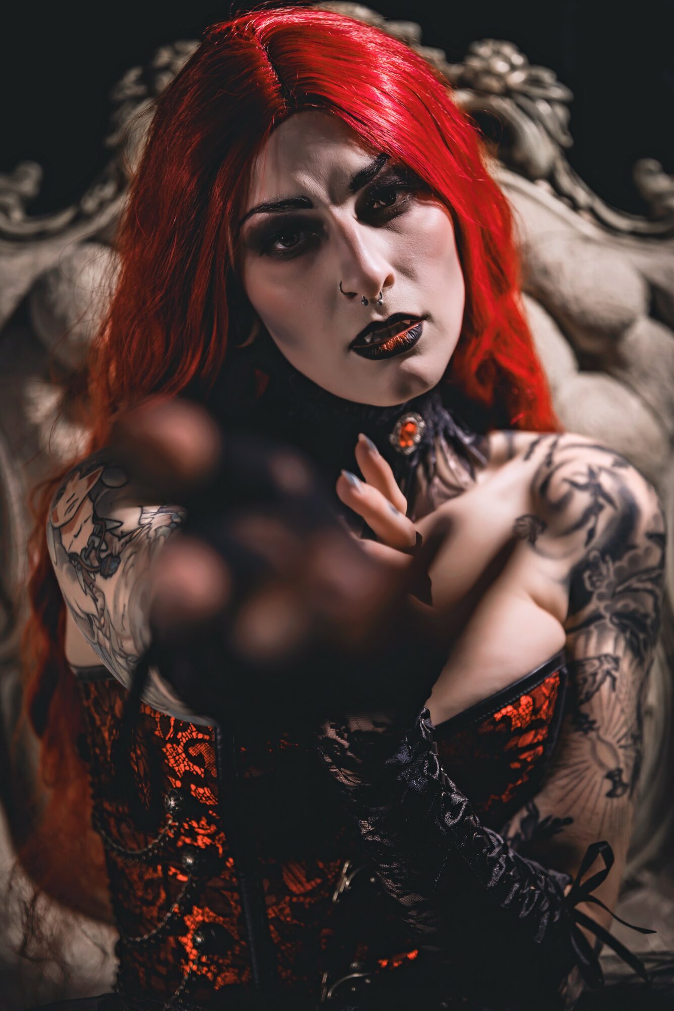 Woman with red hair, dark makeup, tattoos, and gothic clothing sitting on a vintage chair.
