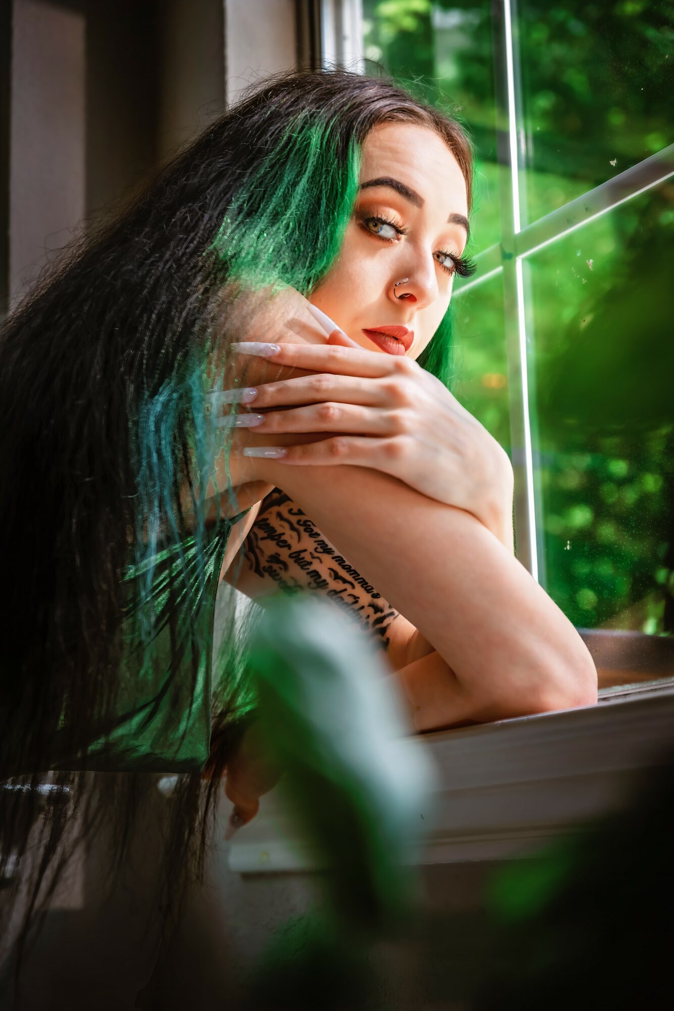 Woman with long black hair and green streaks looking out a window, resting her chin on her hand, with greenery outside.
