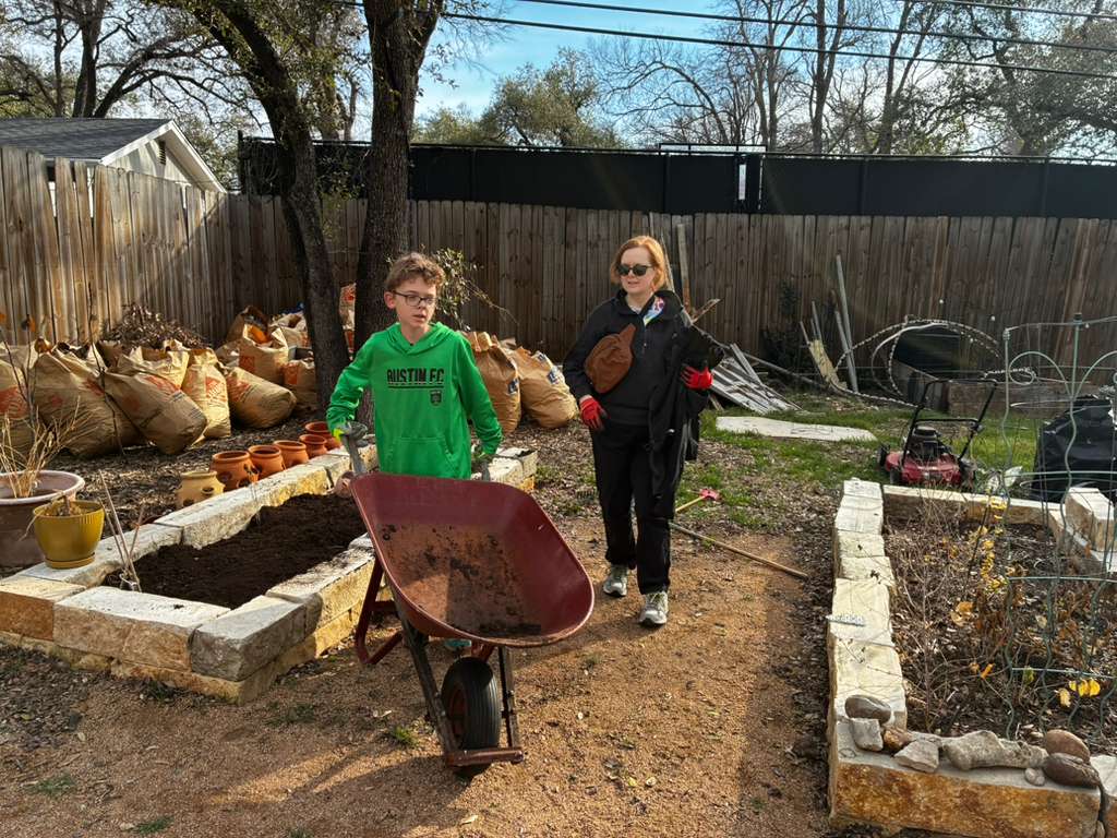 Two children stand in a backyard garden with raised beds, trees, and a wooden fence, one holding a wheelbarrow.
