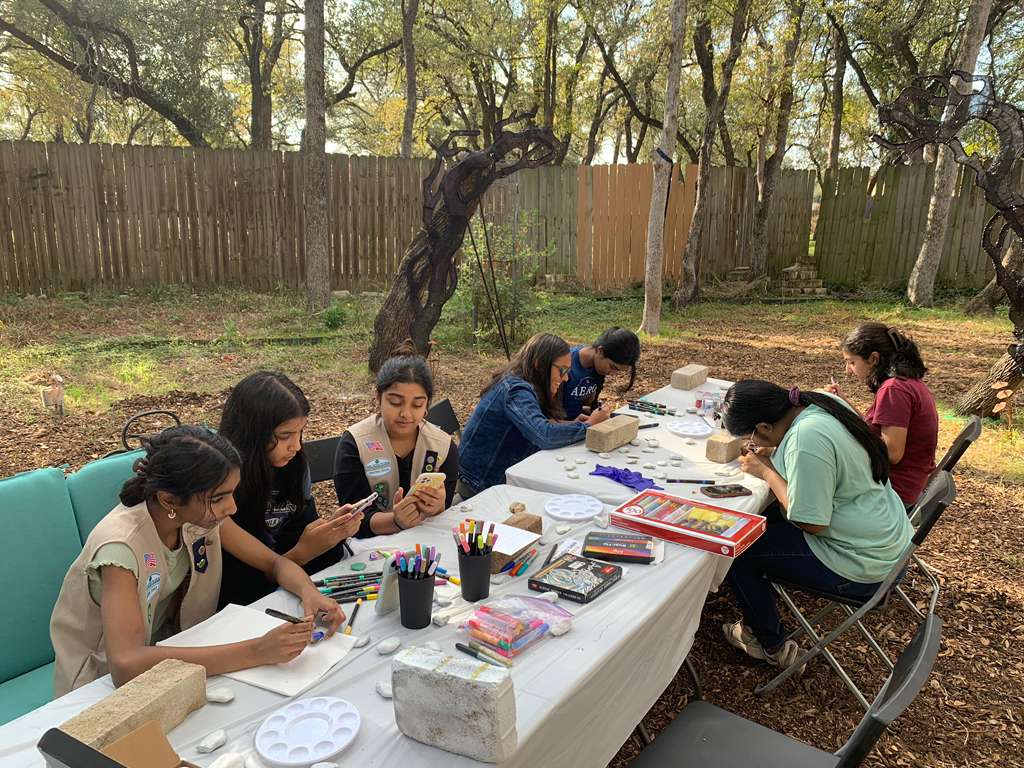 Group of children sitting at a table outdoors, engaging in arts and crafts activities with supplies on the table.