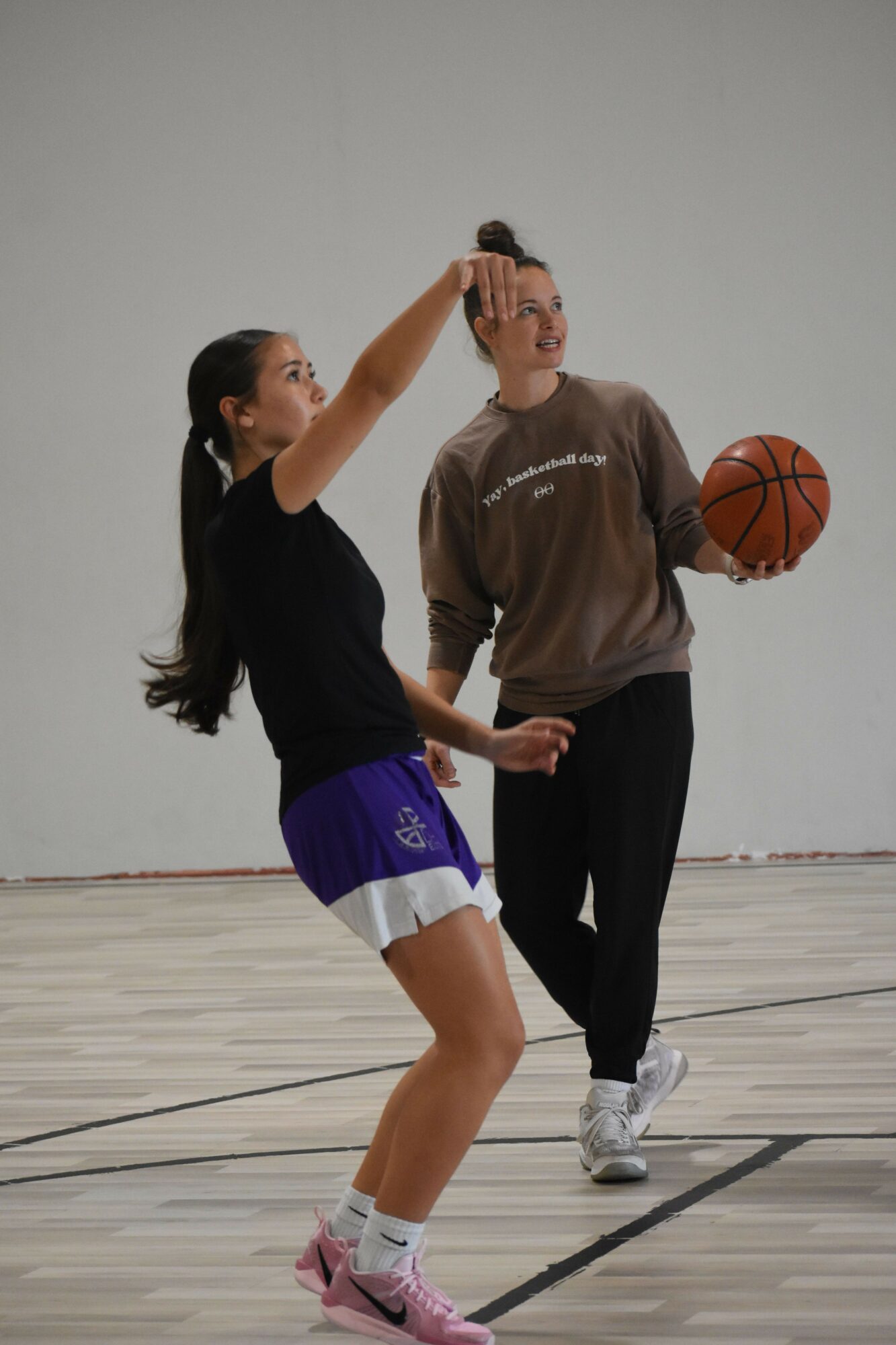 Two women playing basketball indoors, one holding a basketball, the other reaching for it, with a plain wall background.