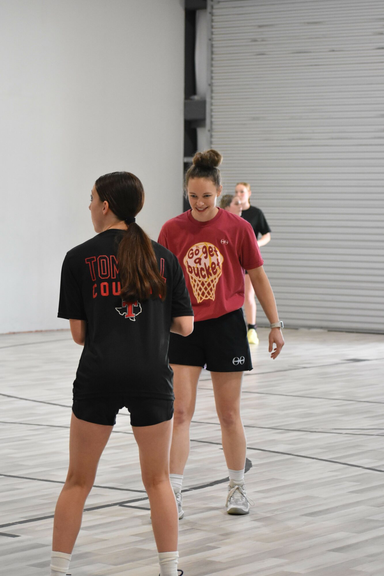 Two women standing and talking indoors, one in a black shirt and shorts, the other in a red shirt and black shorts.