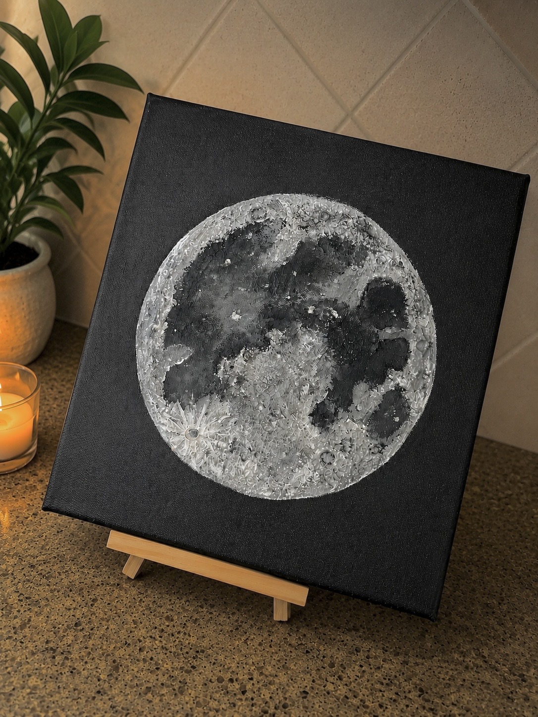 Square canvas with a detailed black and white image of the moon, placed on a small wooden easel on a countertop.