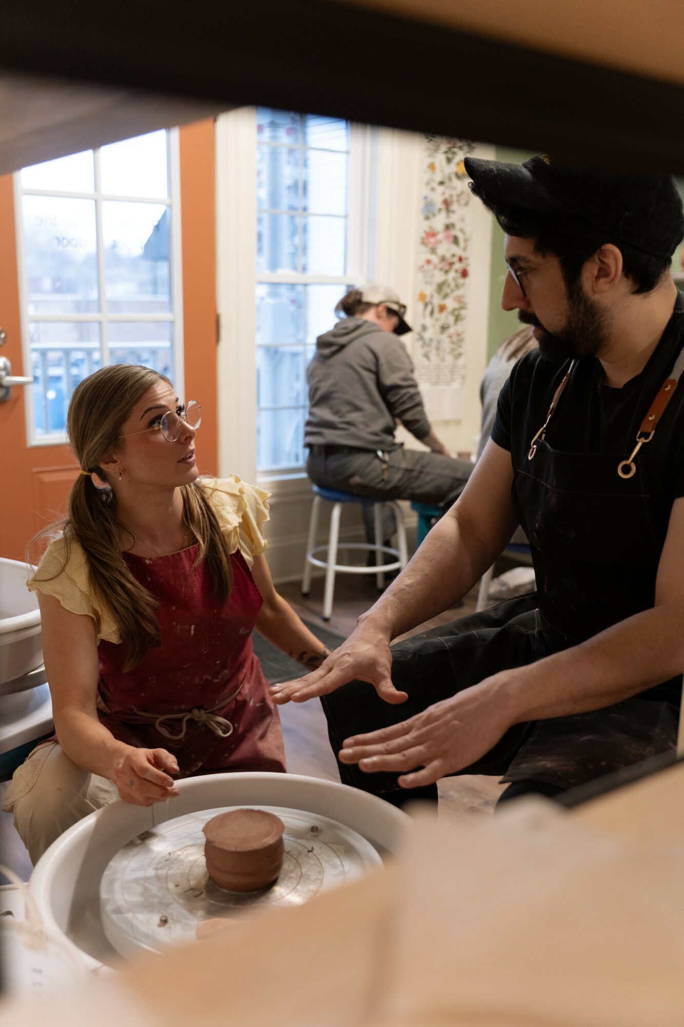 A young girl and a man are in a bakery, with the girl wearing glasses and a red apron, and the man wearing a black shirt. They are interacting near a cake on a table.