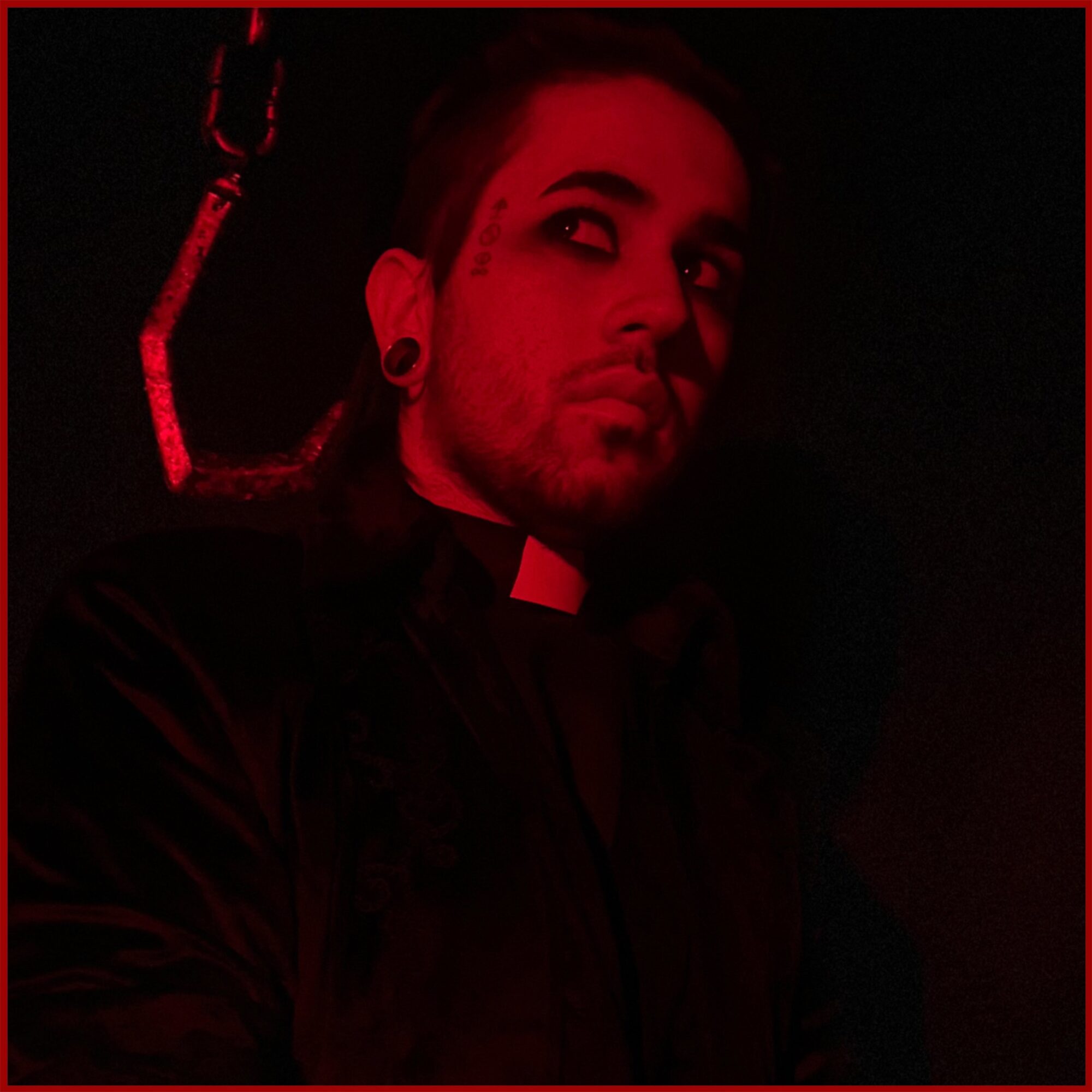 Person with dark hair and makeup, wearing a black outfit, in a dark setting with red lighting.