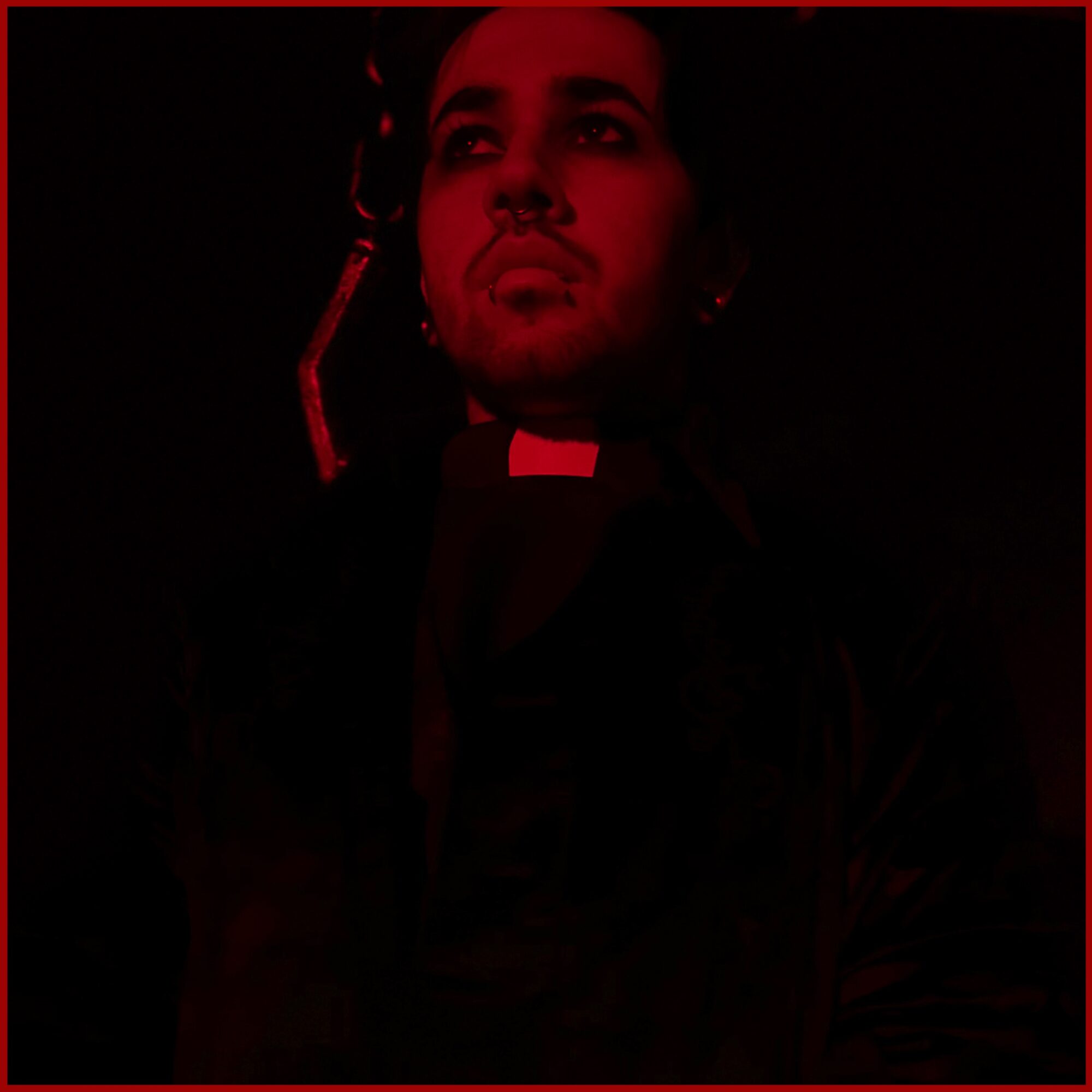 Person with a serious expression, dark background, red lighting, wearing a clerical collar, looking upward.