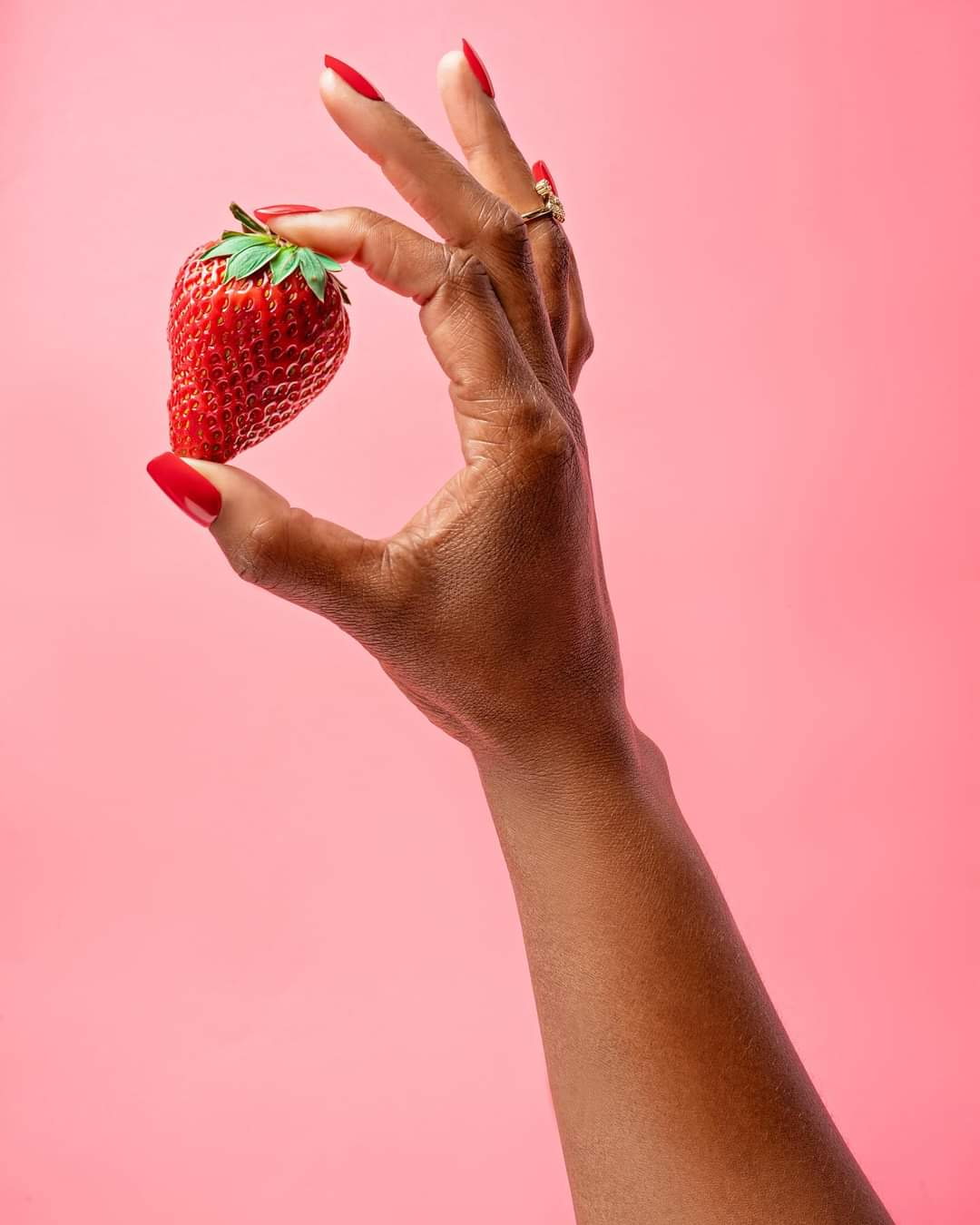 Hand with red nails holding a strawberry against a pink background.