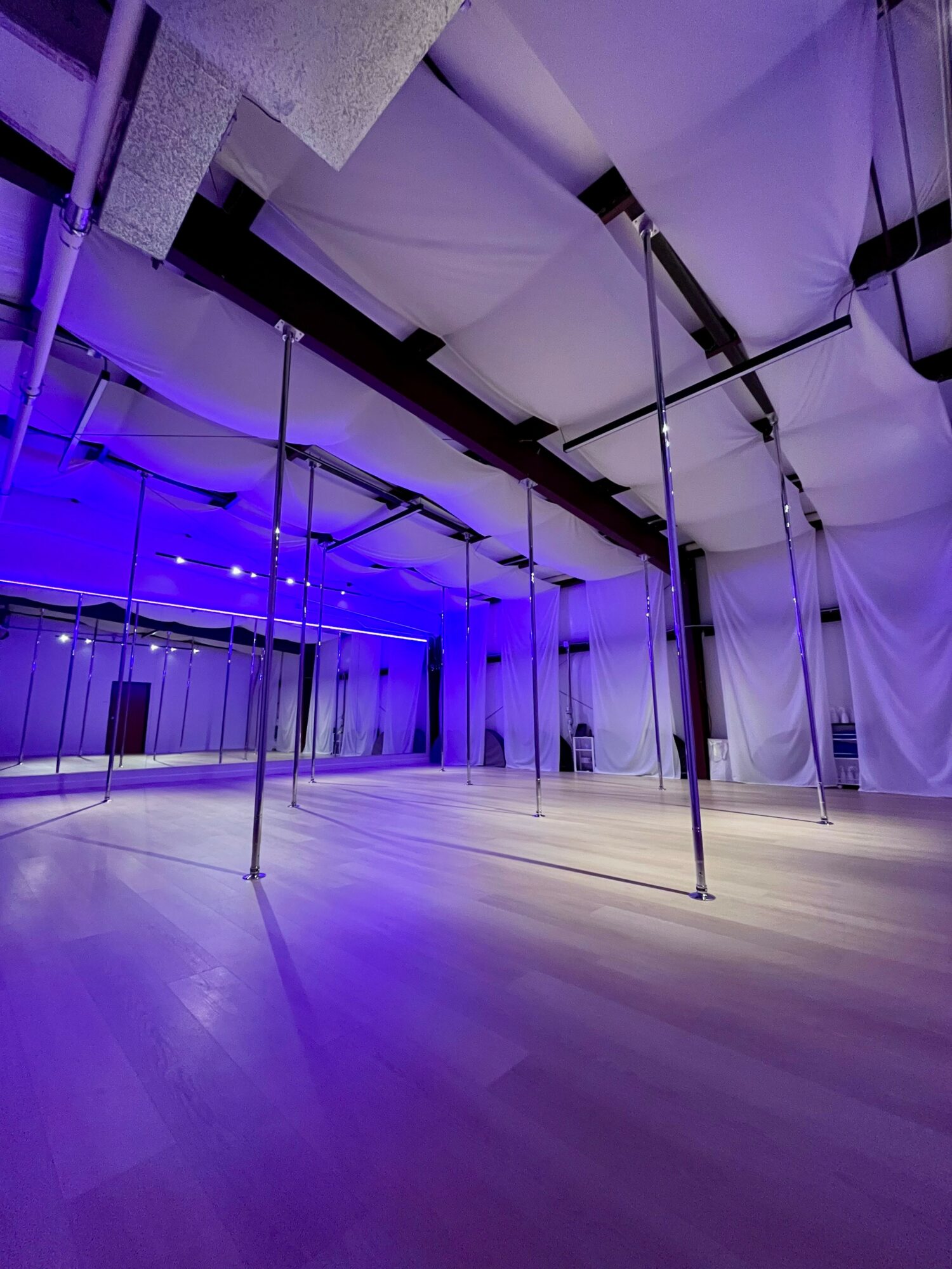 Empty dance studio with poles, purple lighting, and white ceiling panels, wooden floor, and curtains on the walls.