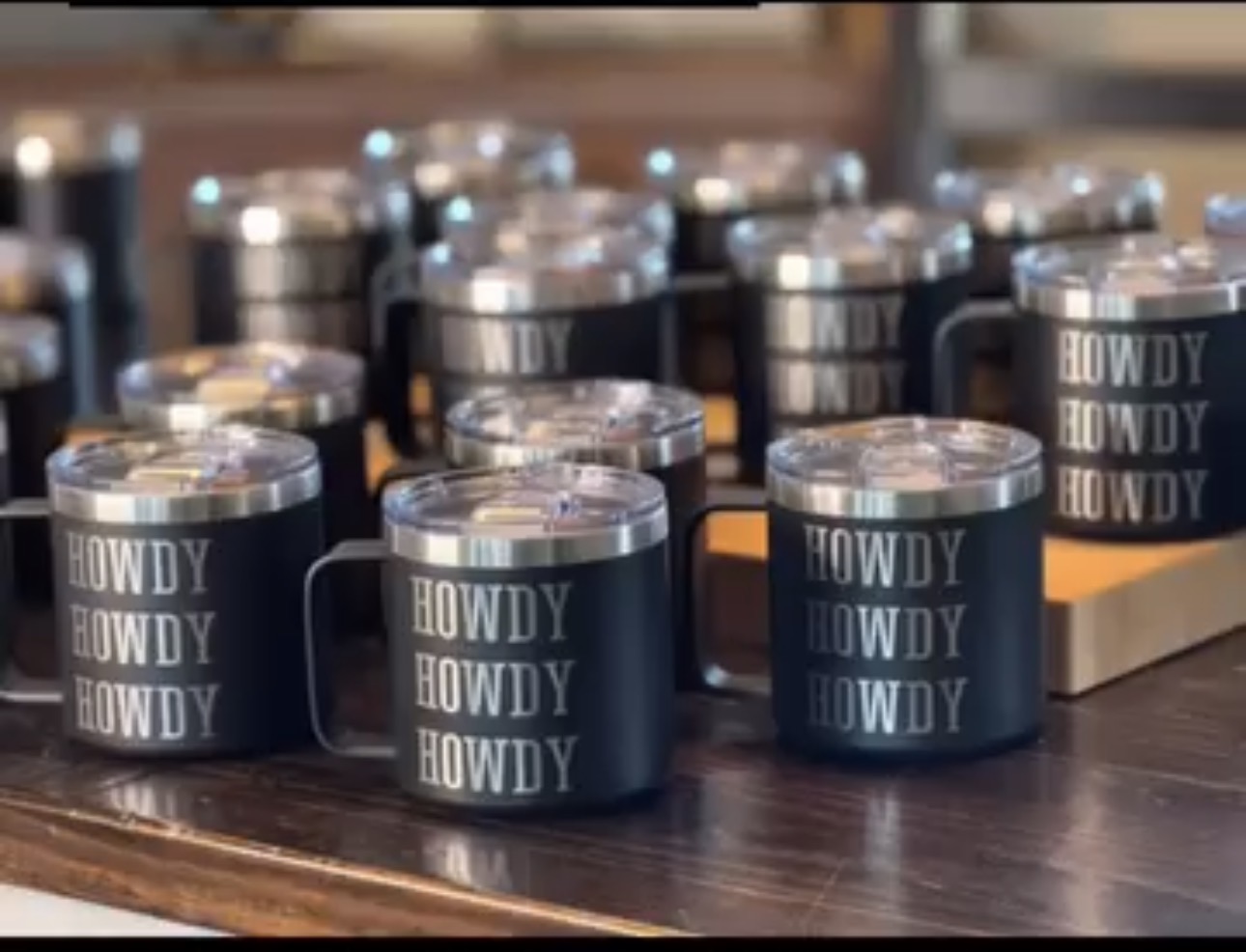 Multiple black mugs with white text saying 'HOWDY' arranged on a wooden surface.