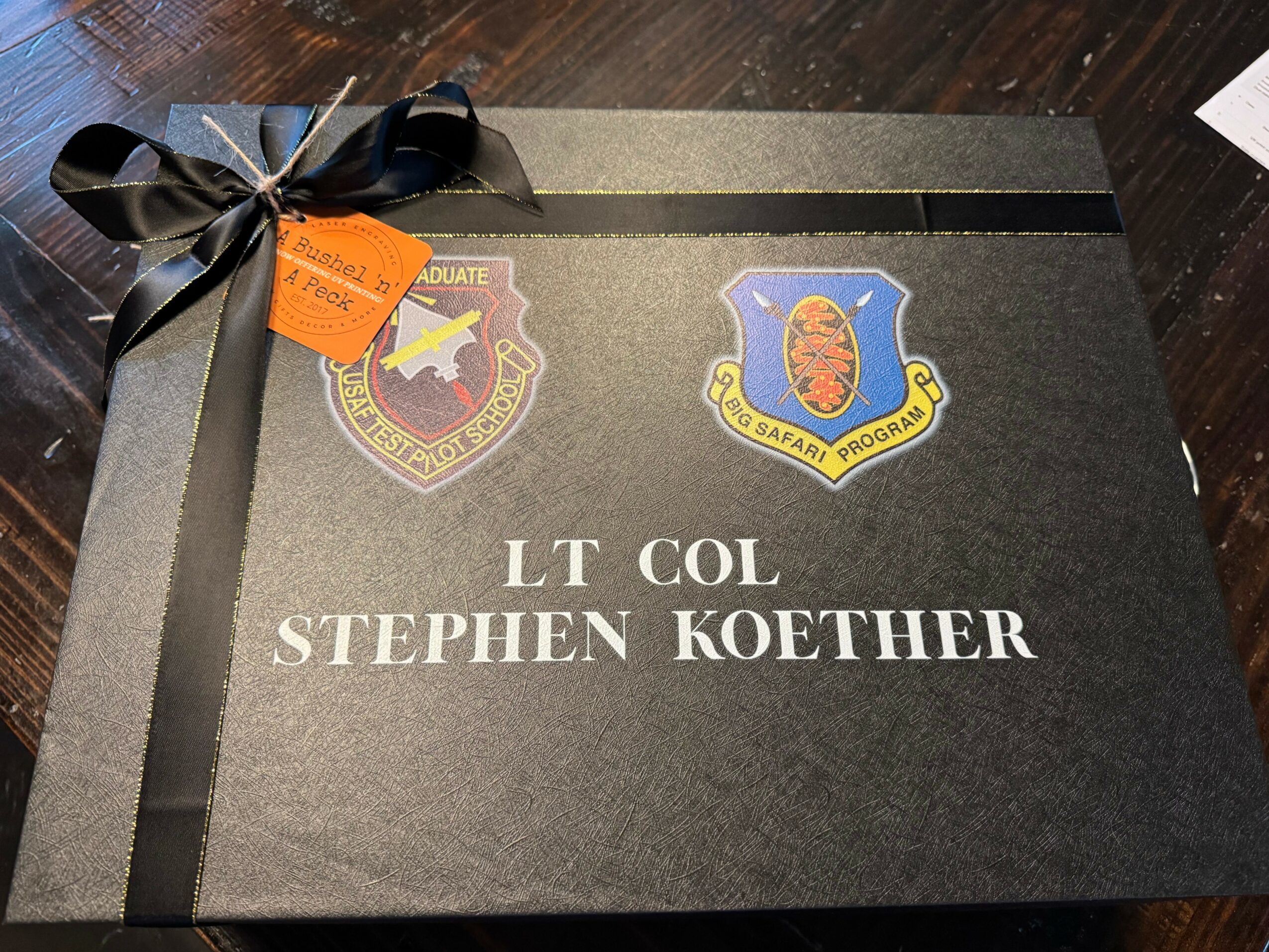 Black gift box with a black ribbon, two patches, and text reading 'LT COL STEPHEN KOETHER' on top.