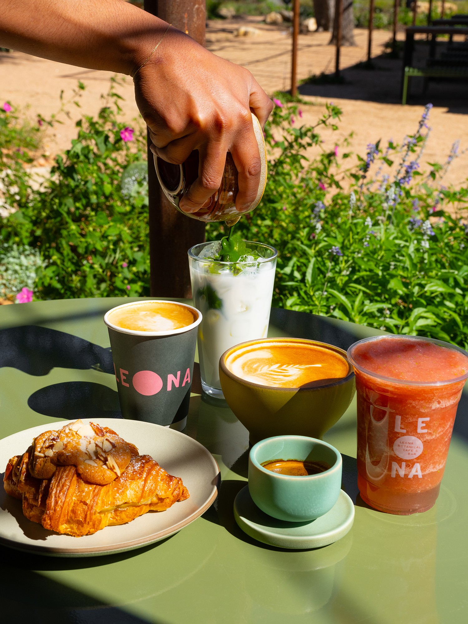 Hand pouring milk into a glass with coffee and croissant on a green table outdoors, surrounded by plants.