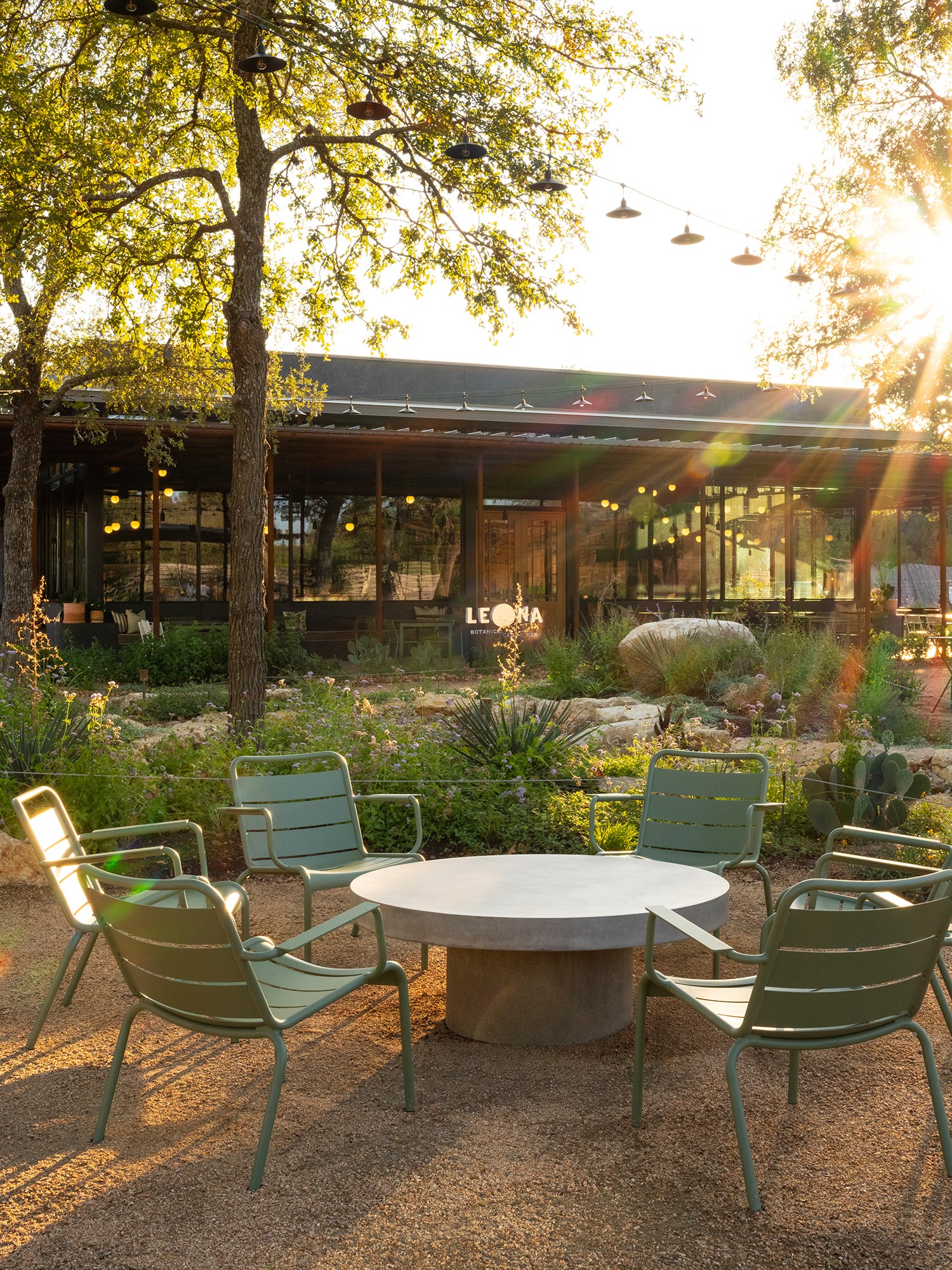 Outdoor seating area with six chairs around a round table, trees, plants, and a building in the background, sunlight shining.