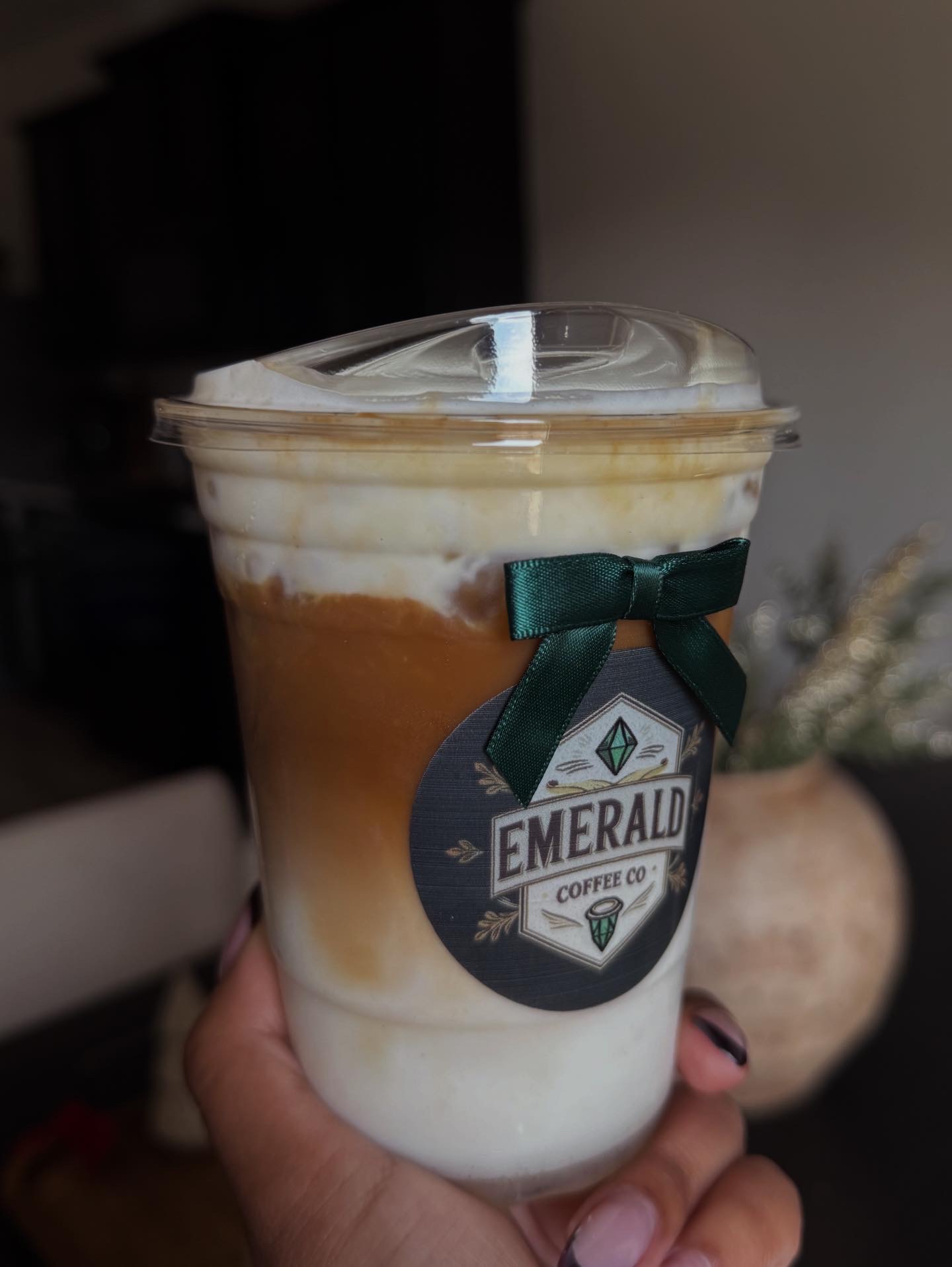 Hand holding a layered iced coffee drink in a clear cup with a lid and a green bow label, against a blurred background.