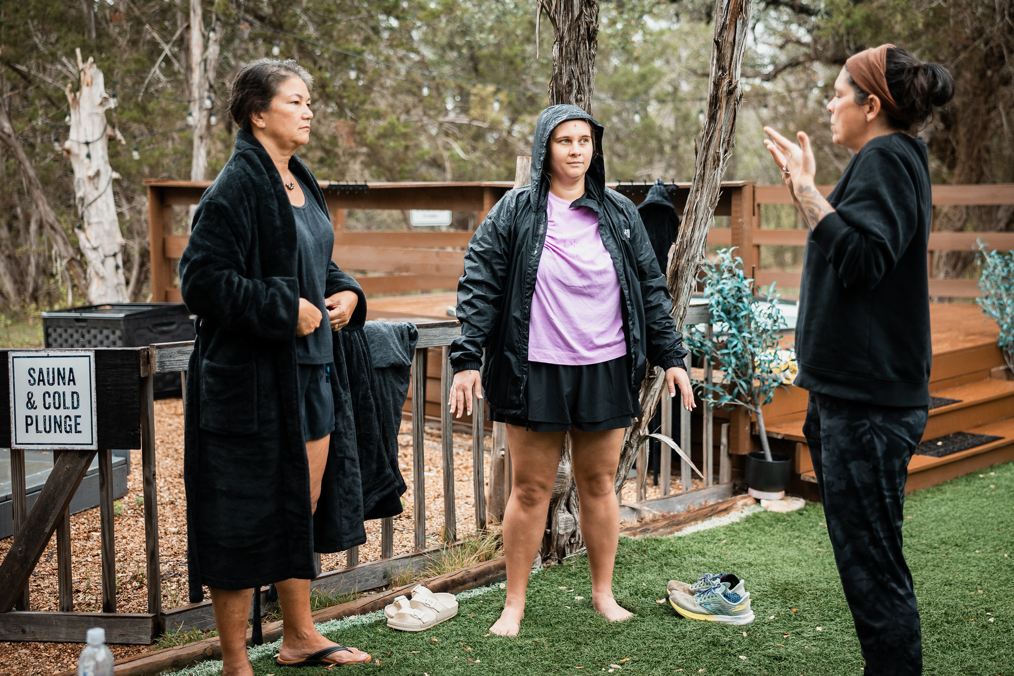 Three women standing outdoors near a wooden fence, engaged in conversation, with trees and a sign in the background.