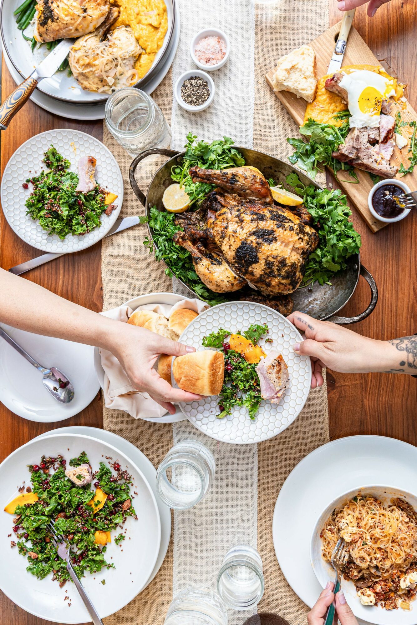 A table with various dishes including roasted chicken, salad, bread, and desserts, with hands serving food. Bright, inviting setting.