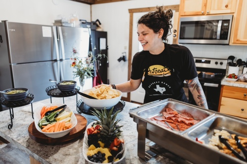 Woman with dark hair in a bun smiling in a kitchen, preparing food on a counter with bowls and trays, refrigerator and microwave in background.
