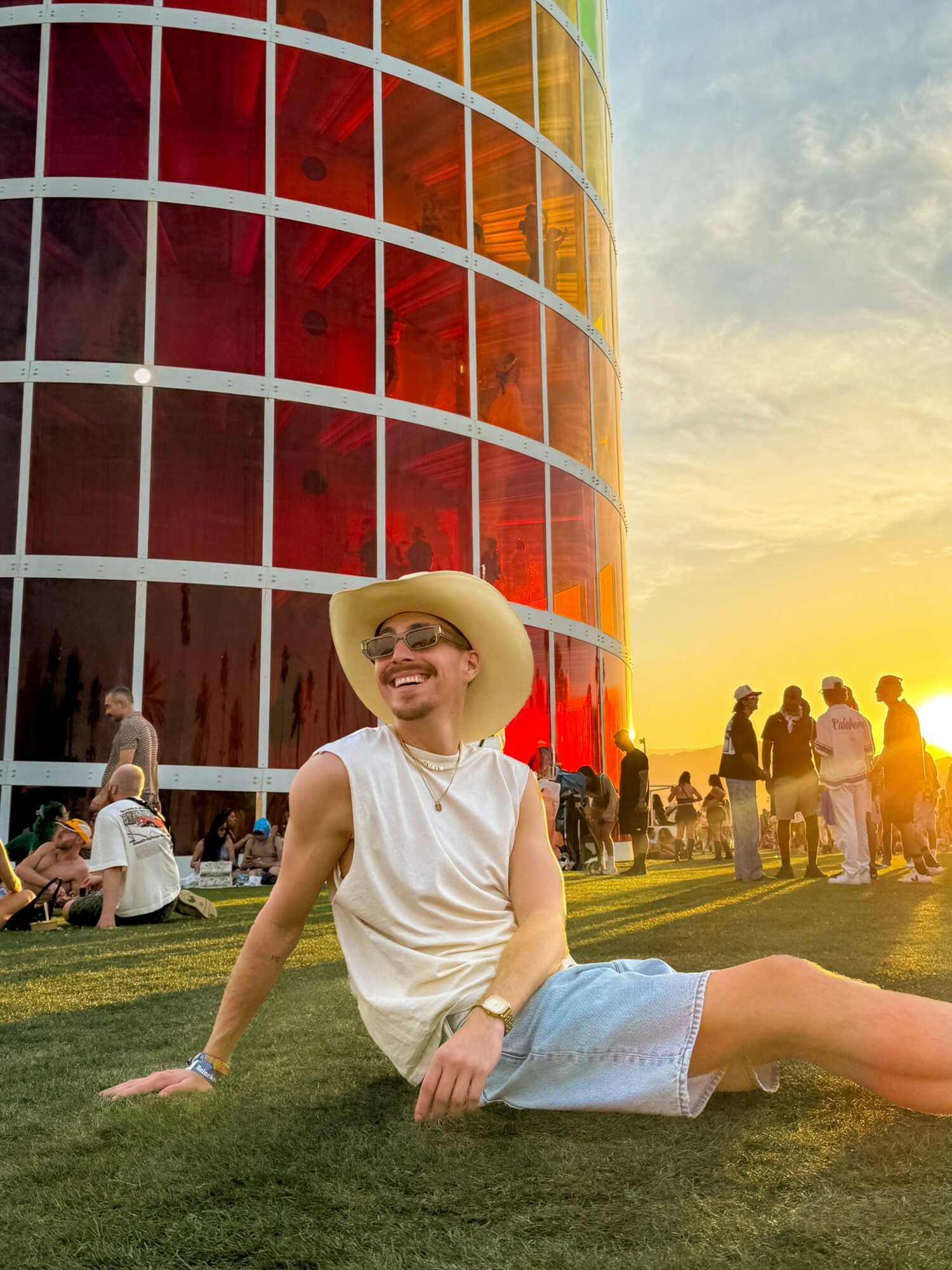 Person sitting on grass in front of a colorful glass building during sunset, smiling, wearing a hat and casual clothes.