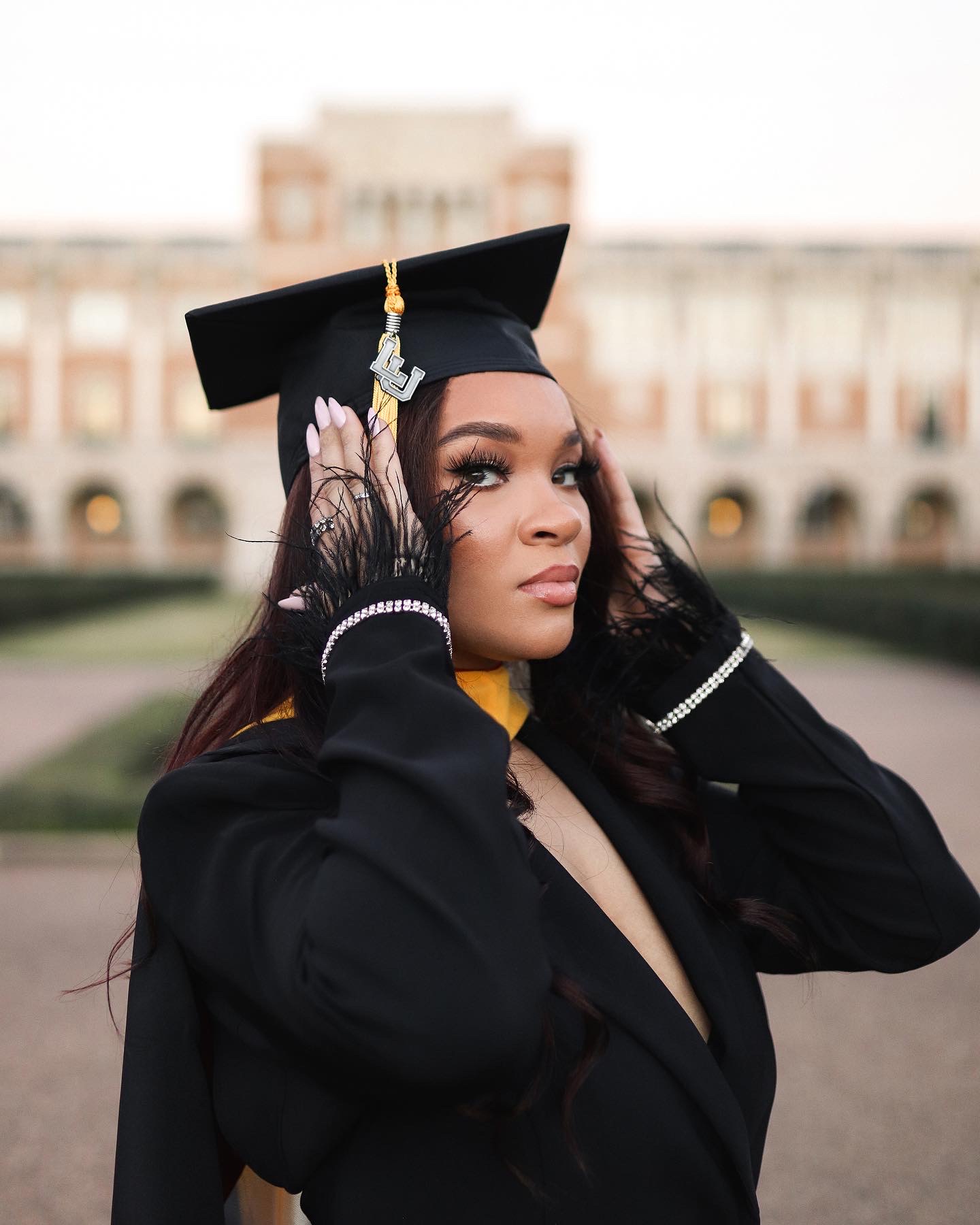 Woman in graduation cap and gown adjusting her cap outdoors, with a large building in the background.