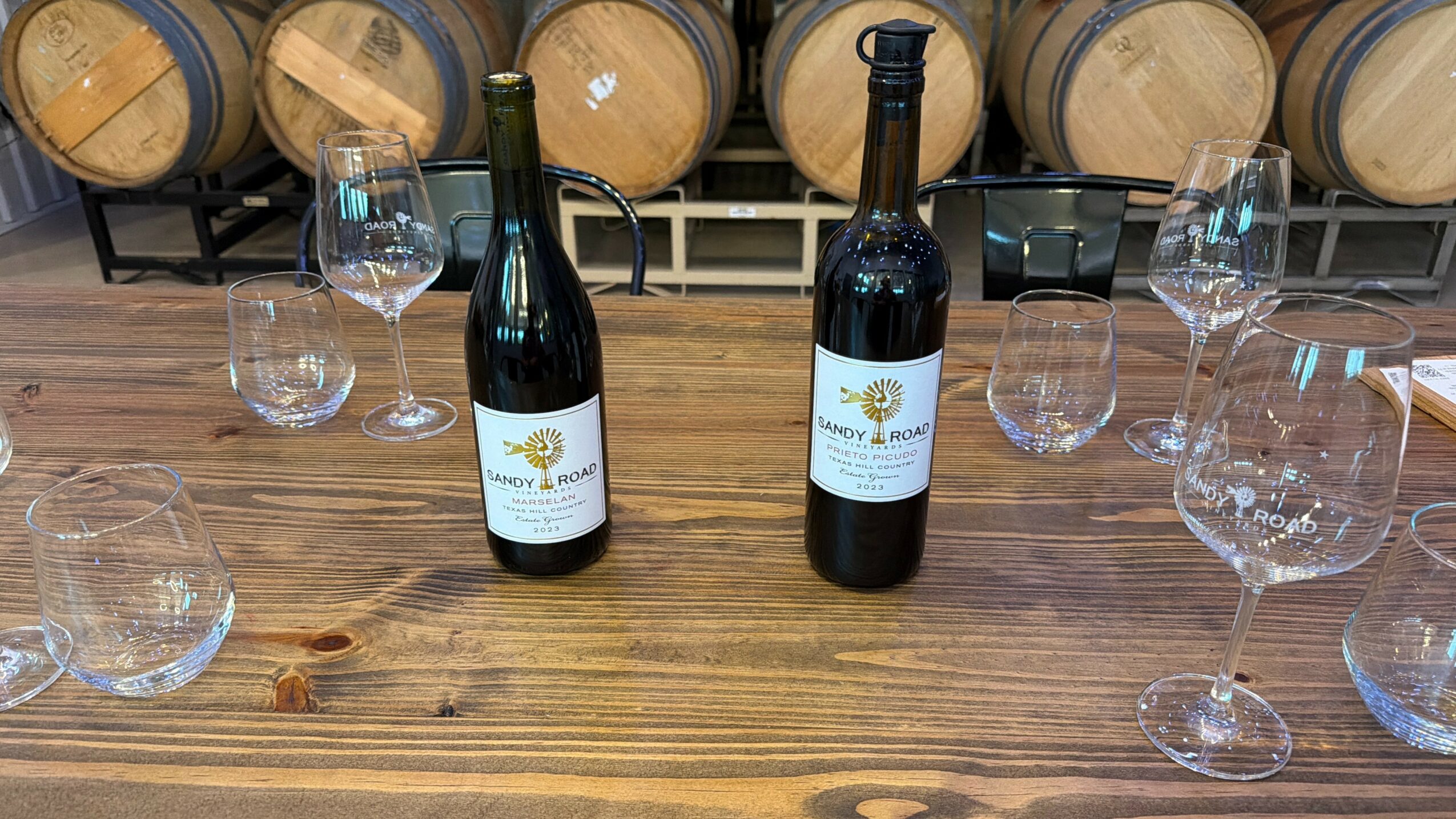 Two bottles of wine on a wooden table with glasses and barrels in the background.
