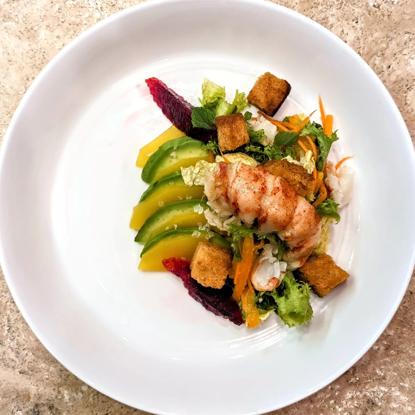 Plate with sliced avocado, cooked lobster, croutons, and colorful vegetables on a white dish.